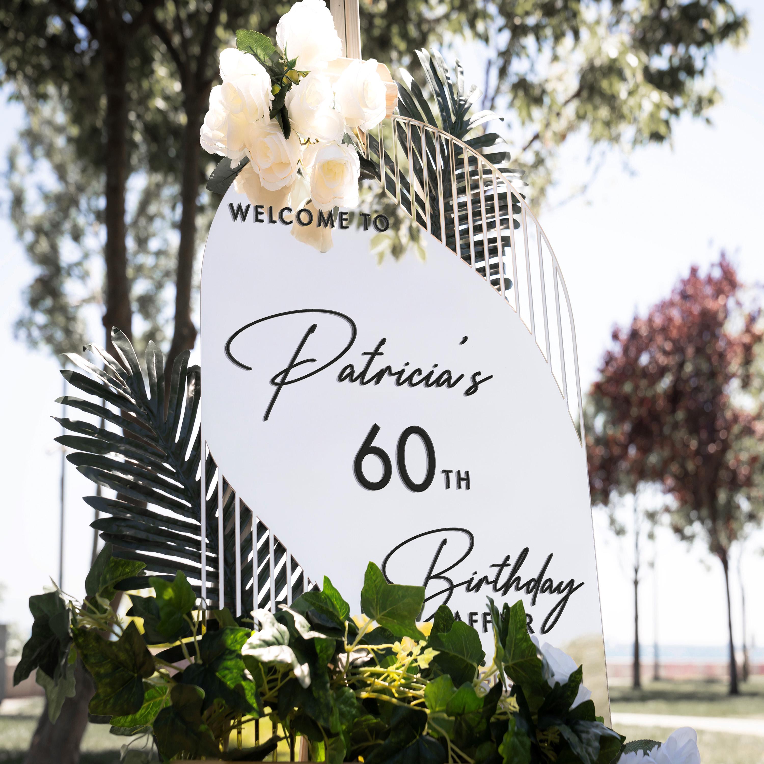 A white sign with black text welcoming guests to Patricia's 60th birthday party, surrounded by greenery and flowers.
