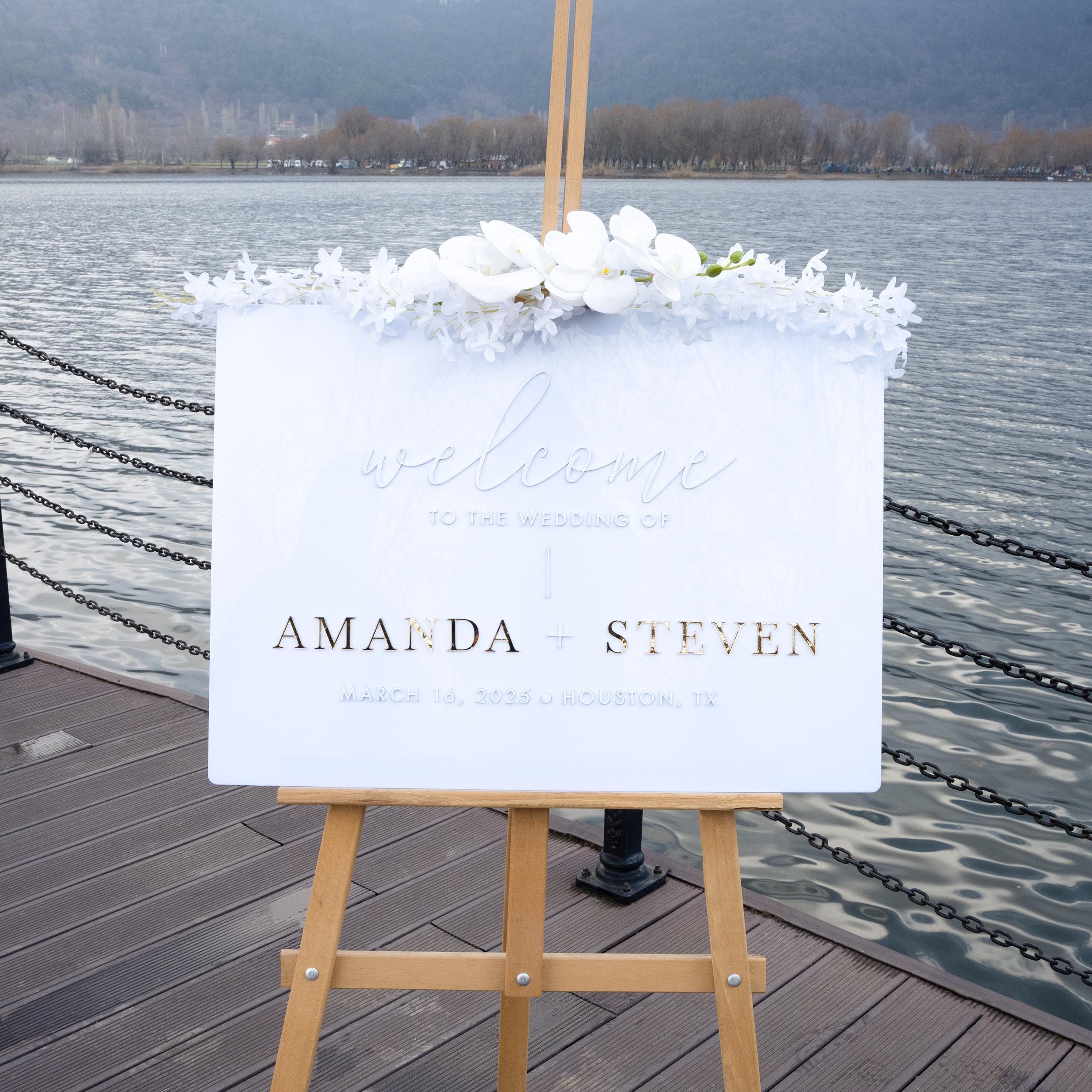 Minimalist white acrylic wedding welcome sign with elegant gold 3D lettering, featuring the couple’s names and wedding details. The clean and sophisticated design adds a timeless and modern touch to any wedding entrance.