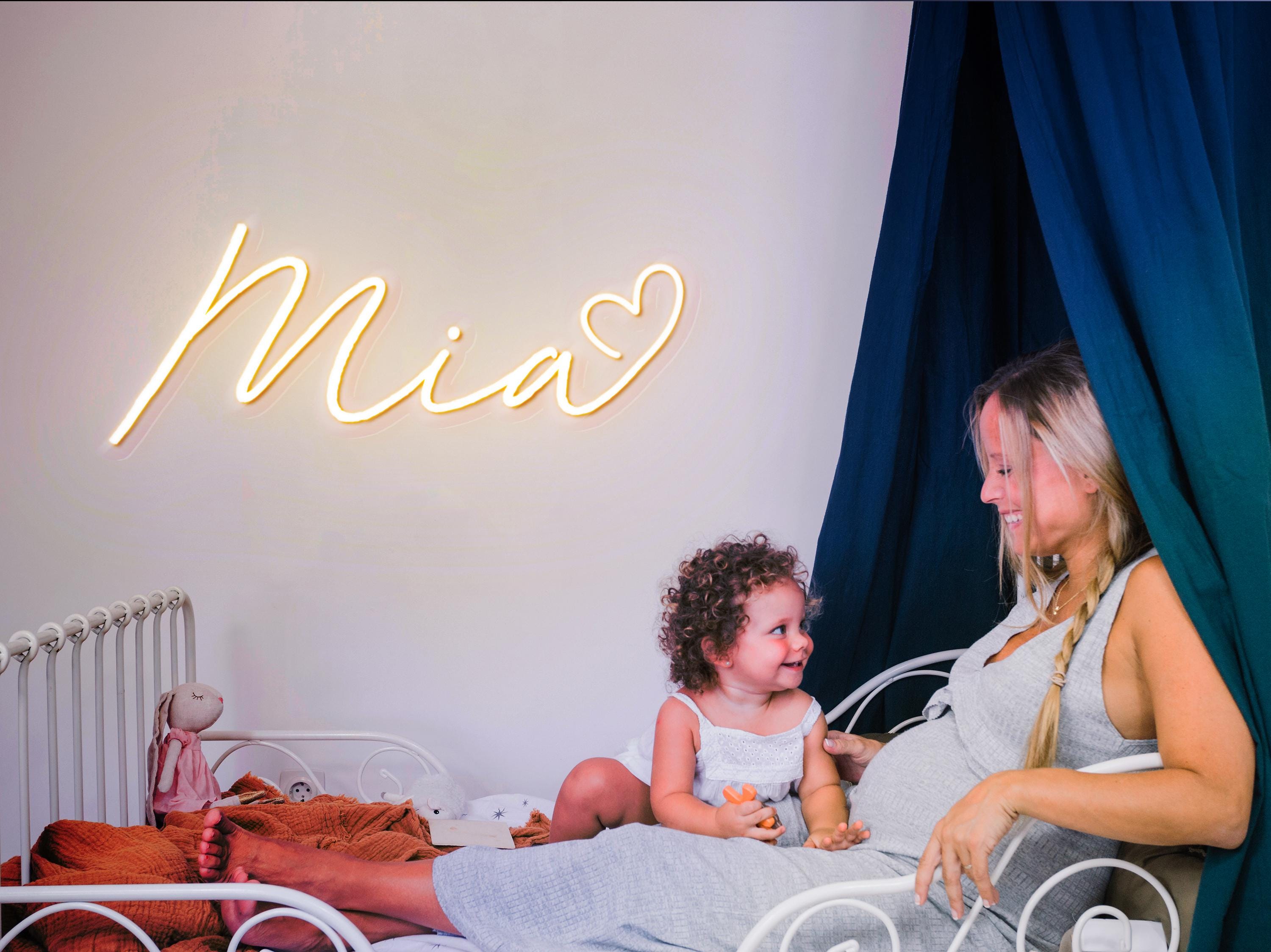 A pregnant woman and her young daughter are sitting together on a bed, with a neon sign that reads "Mia" in the background.