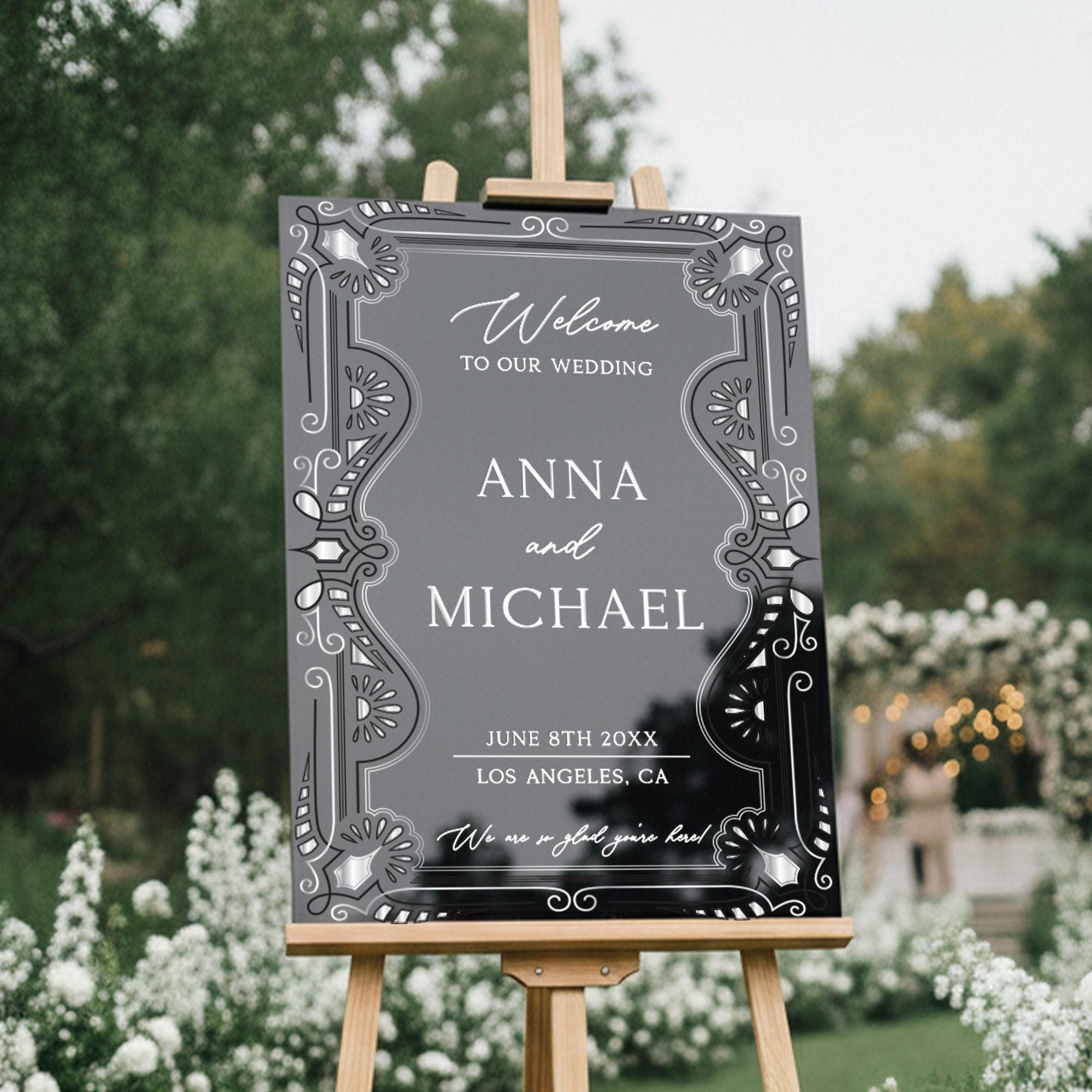 a black and white wedding welcome sign with decorative elements, standing in a garden setting with trees and flowers in the background.