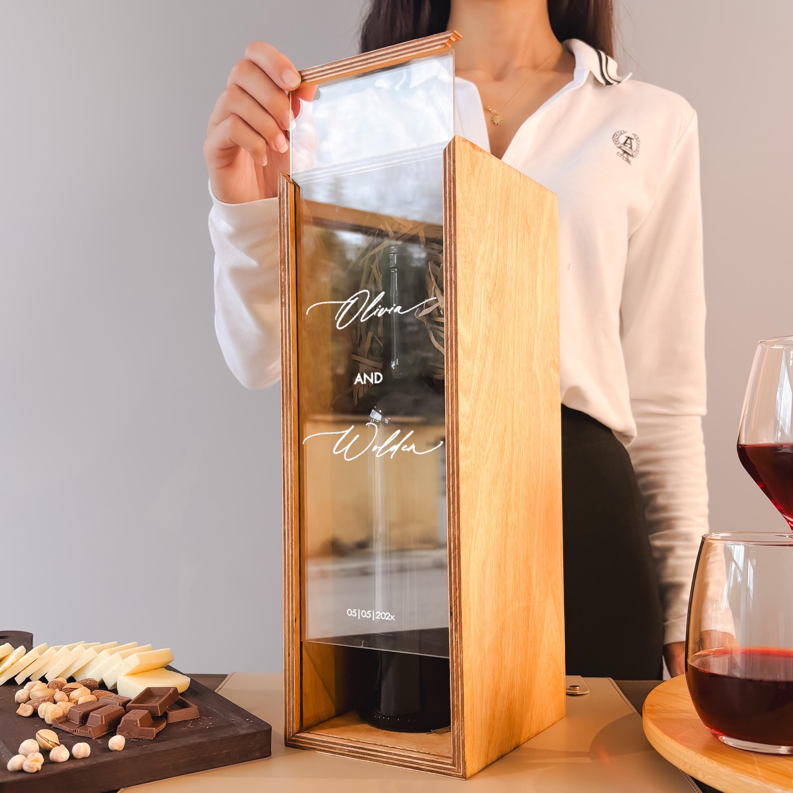 A person is holding a wooden wine box with a glass of red wine inside it, and there are other items on a table in the background.