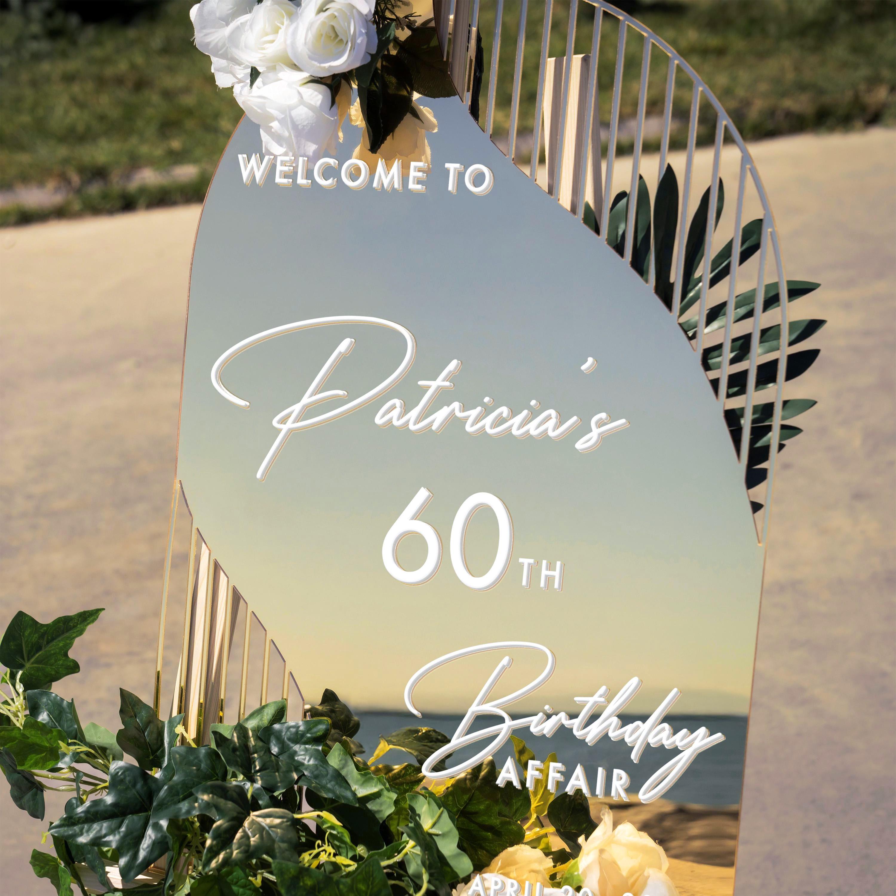 a sign that reads "Welcome to Patricia's 60th Birthday Affair" with a mirror and some greenery as decoration.