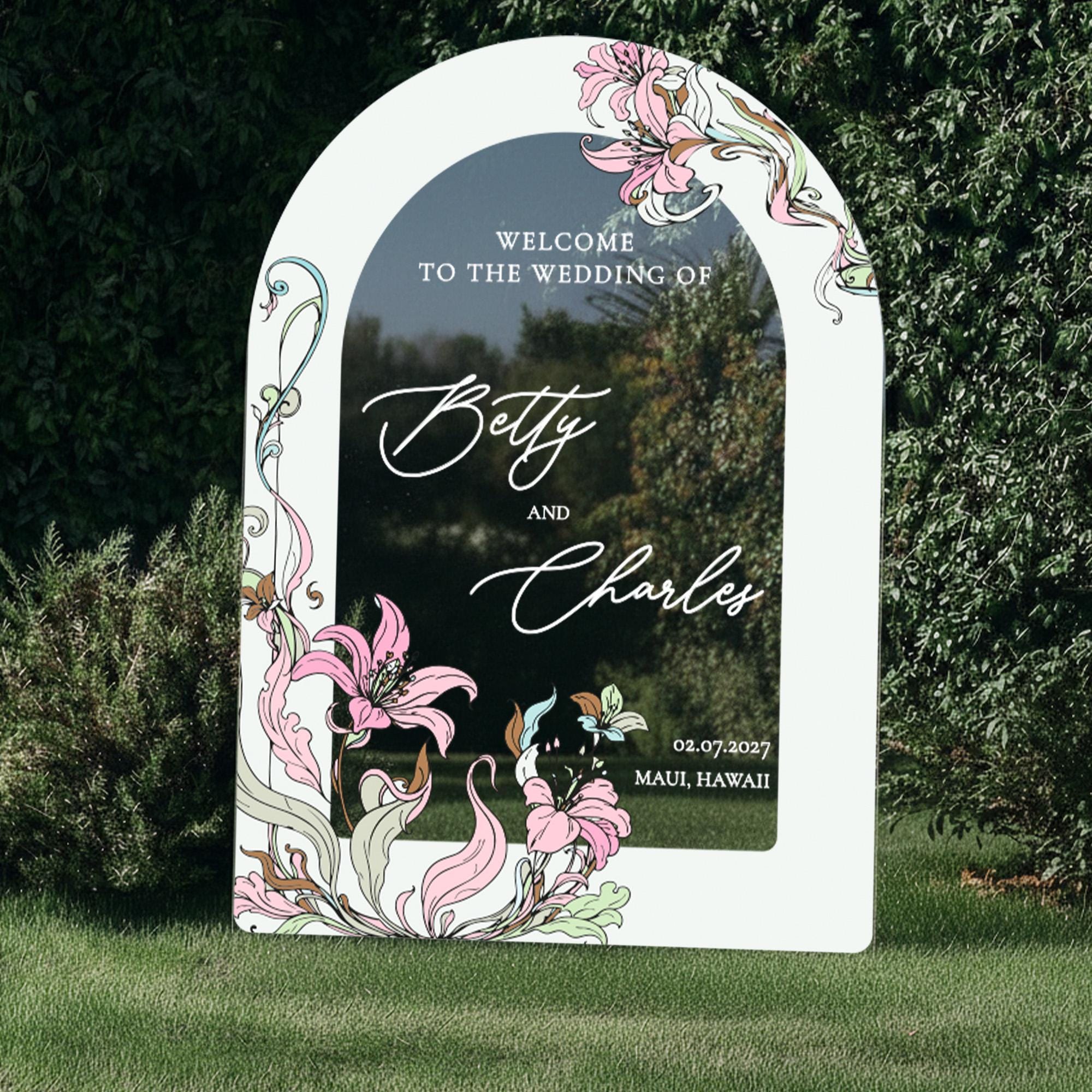 a white sign with floral designs and text, standing in a grassy area surrounded by trees.