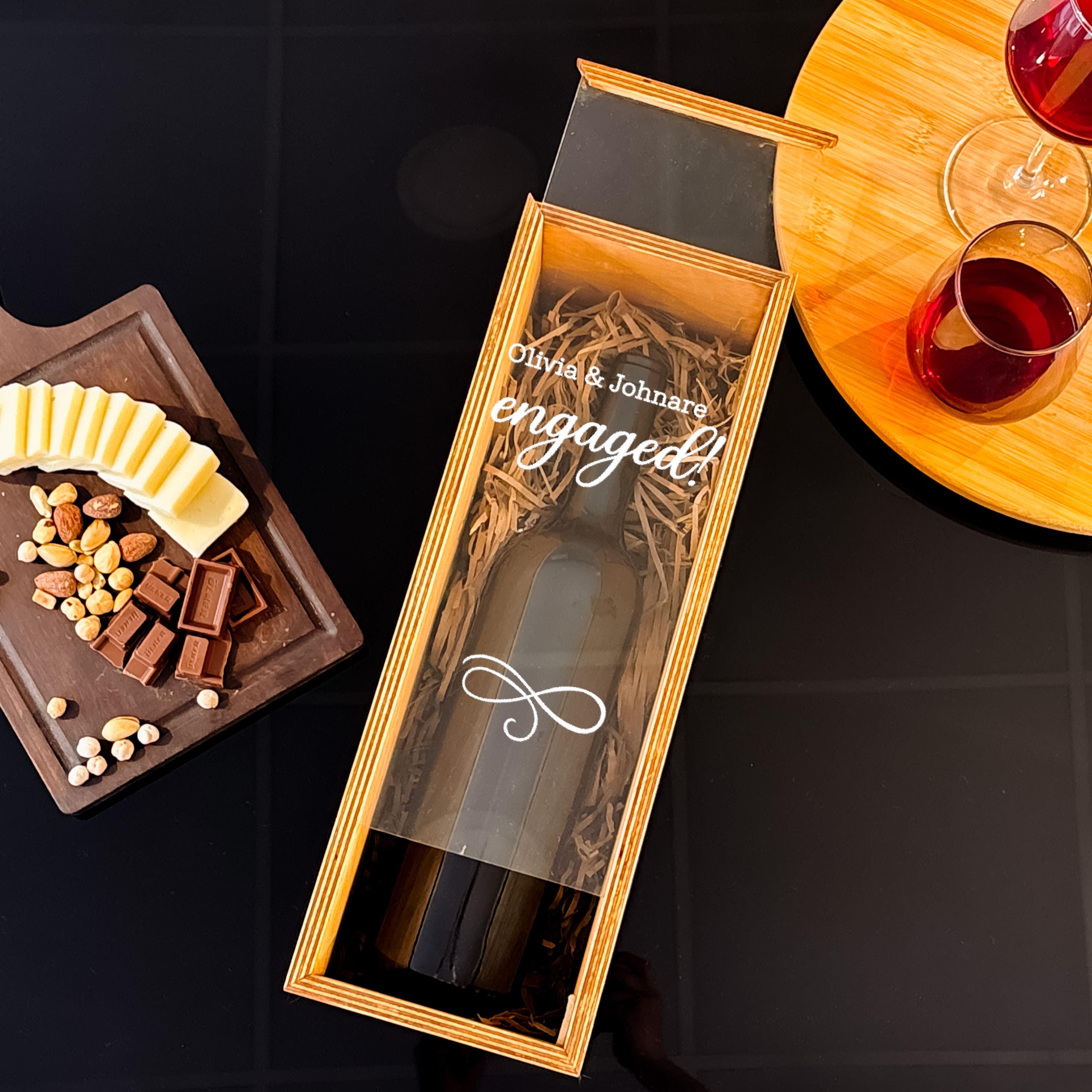 A wooden box containing a bottle of wine, with a label that reads "Olivia & Johnathan engaged", along with a cheese platter and glasses of red wine on a wooden board.