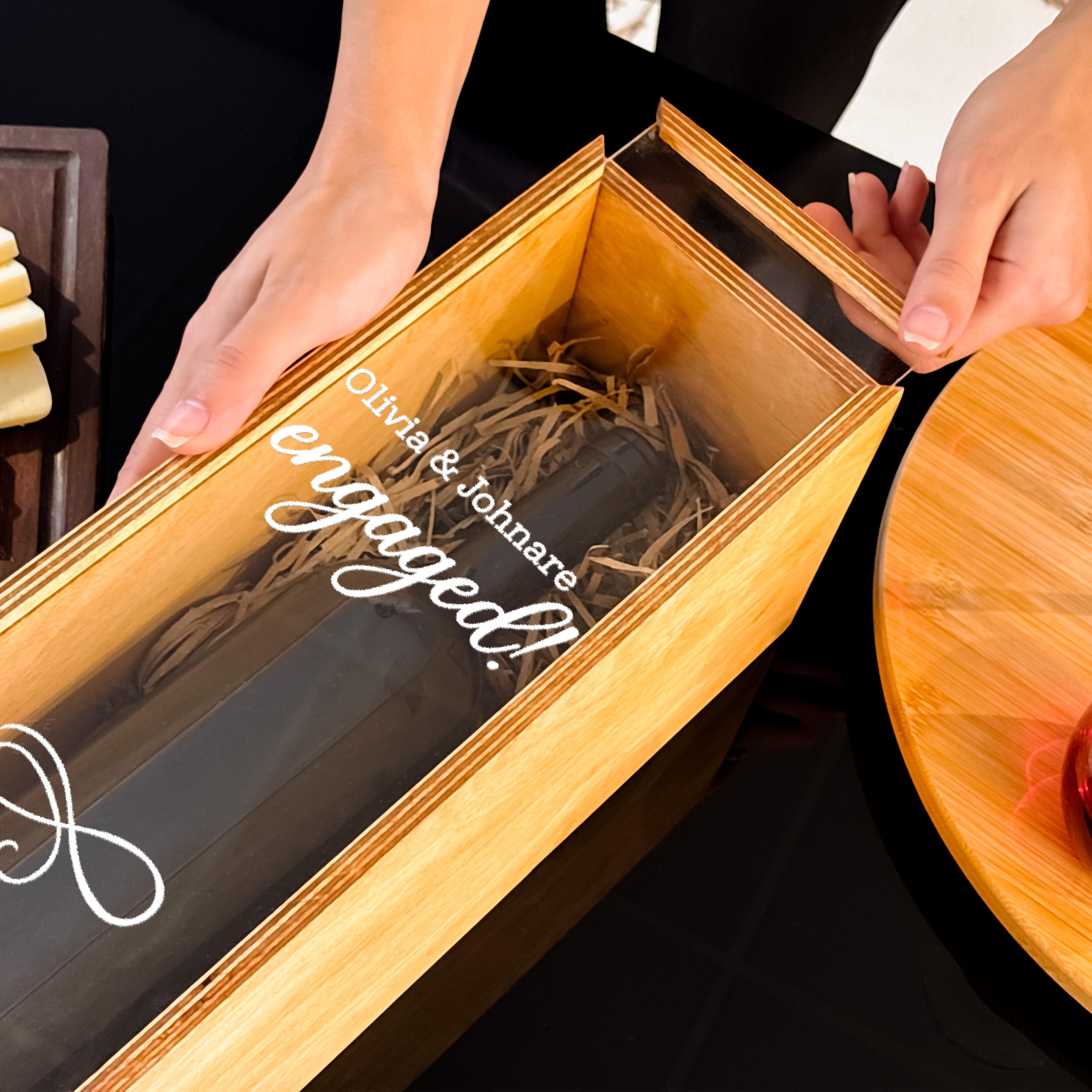 A person's hands are holding a wooden box with a clear plastic window, which contains a bottle of wine and some shredded wood.