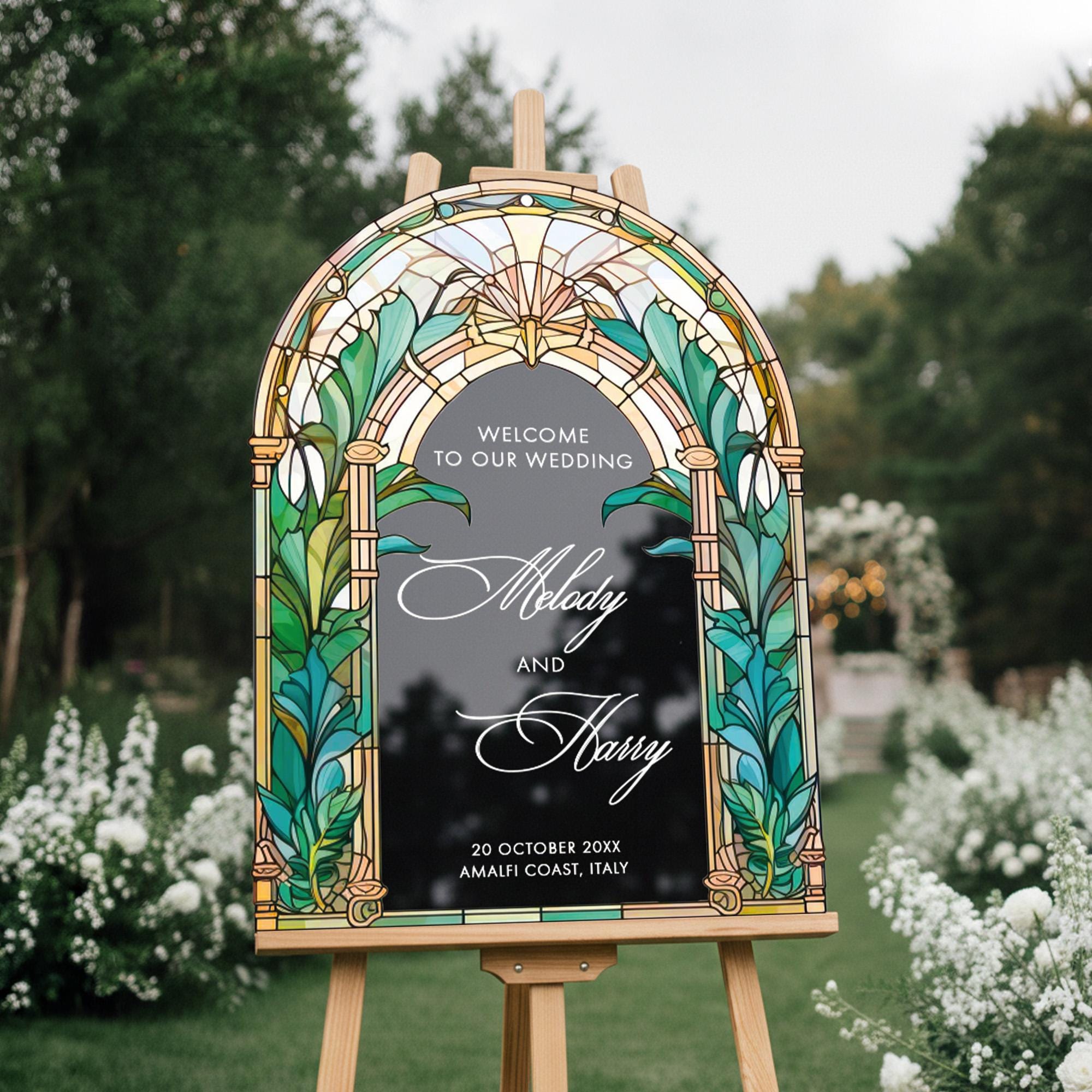 a wooden wedding sign with a stained glass design, standing in a garden setting with white flowers in the background.