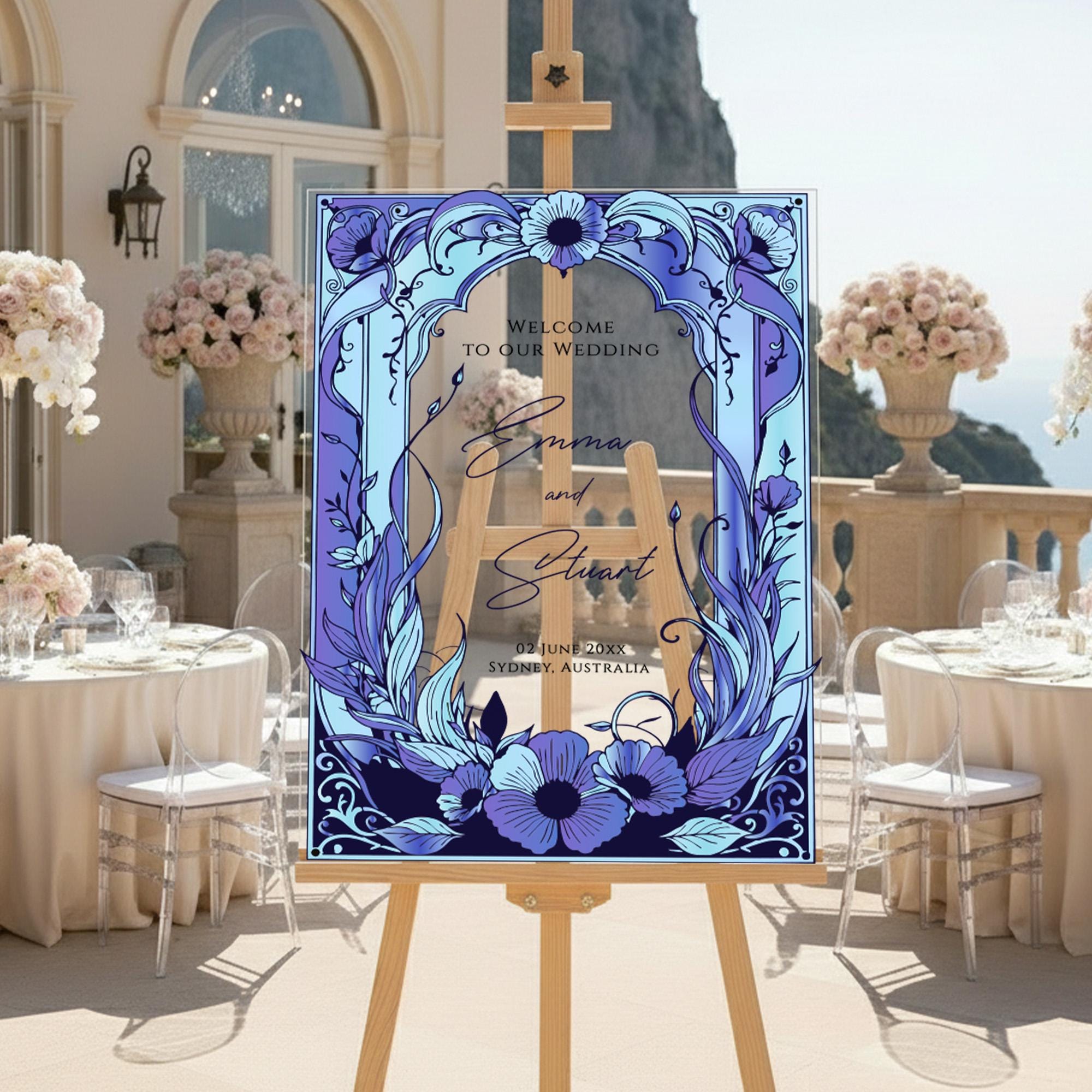 a wedding invitation with a blue and purple floral design, displayed on an easel in front of a table with white tablecloths and floral centerpieces.