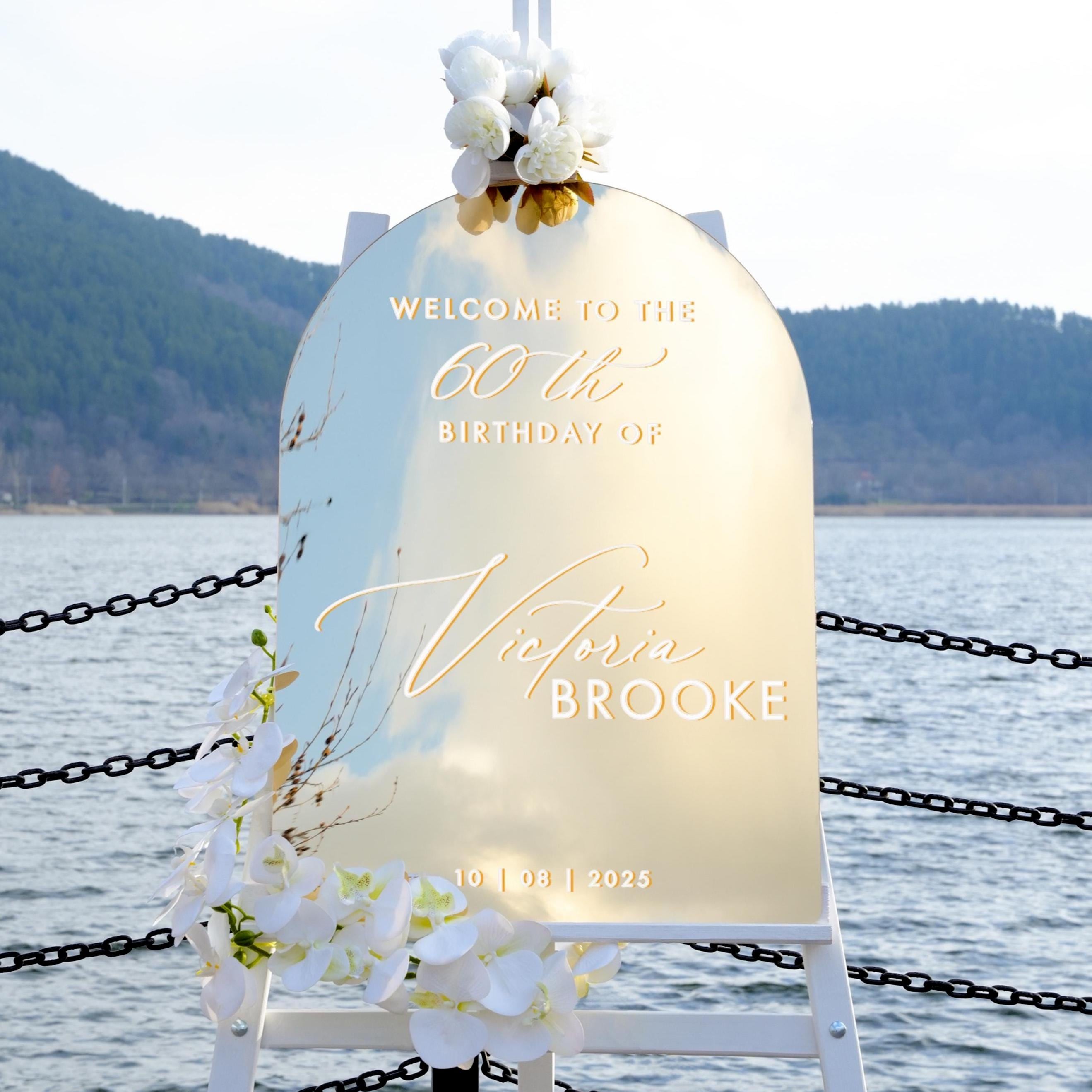 a sign welcoming guests to the 60th birthday of Victoria Brooke, with a scenic backdrop of a body of water and mountains.