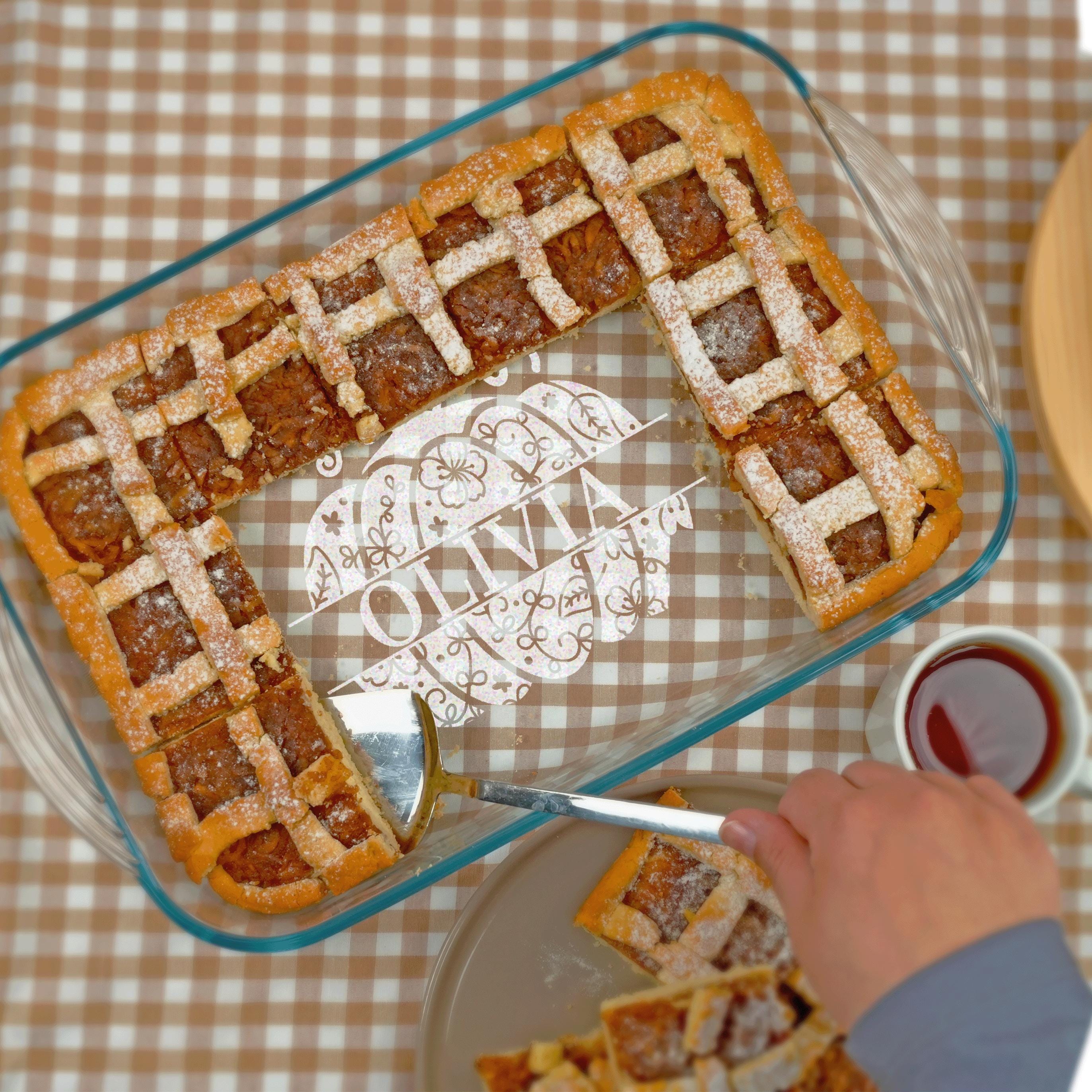 A glass baking dish filled with a dessert, containing a pastry with a lattice crust and powdered sugar, is placed on a table with a checkered tablecloth. A person's hand is reaching towards the dessert, and there is a cup of tea nearby.