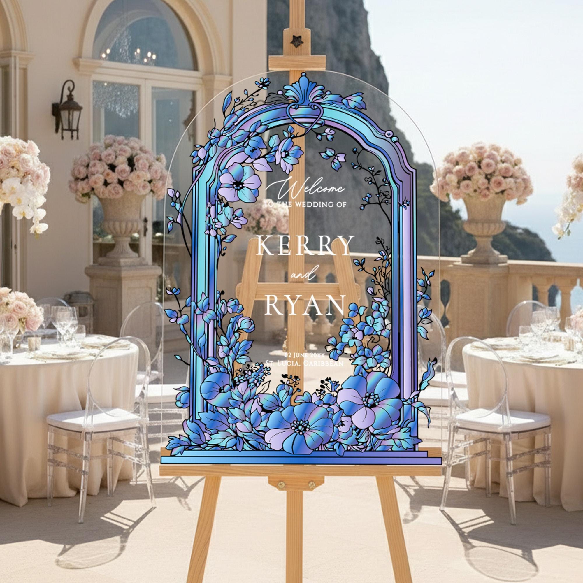 a wedding reception setup with a large, colorful sign displaying the names 'Kerry' and 'Ryan' in a floral design. The sign is placed in front of a table with white tablecloths and clear glass chairs. The background includes a scenic view of the ocean and a building with arched windows.
