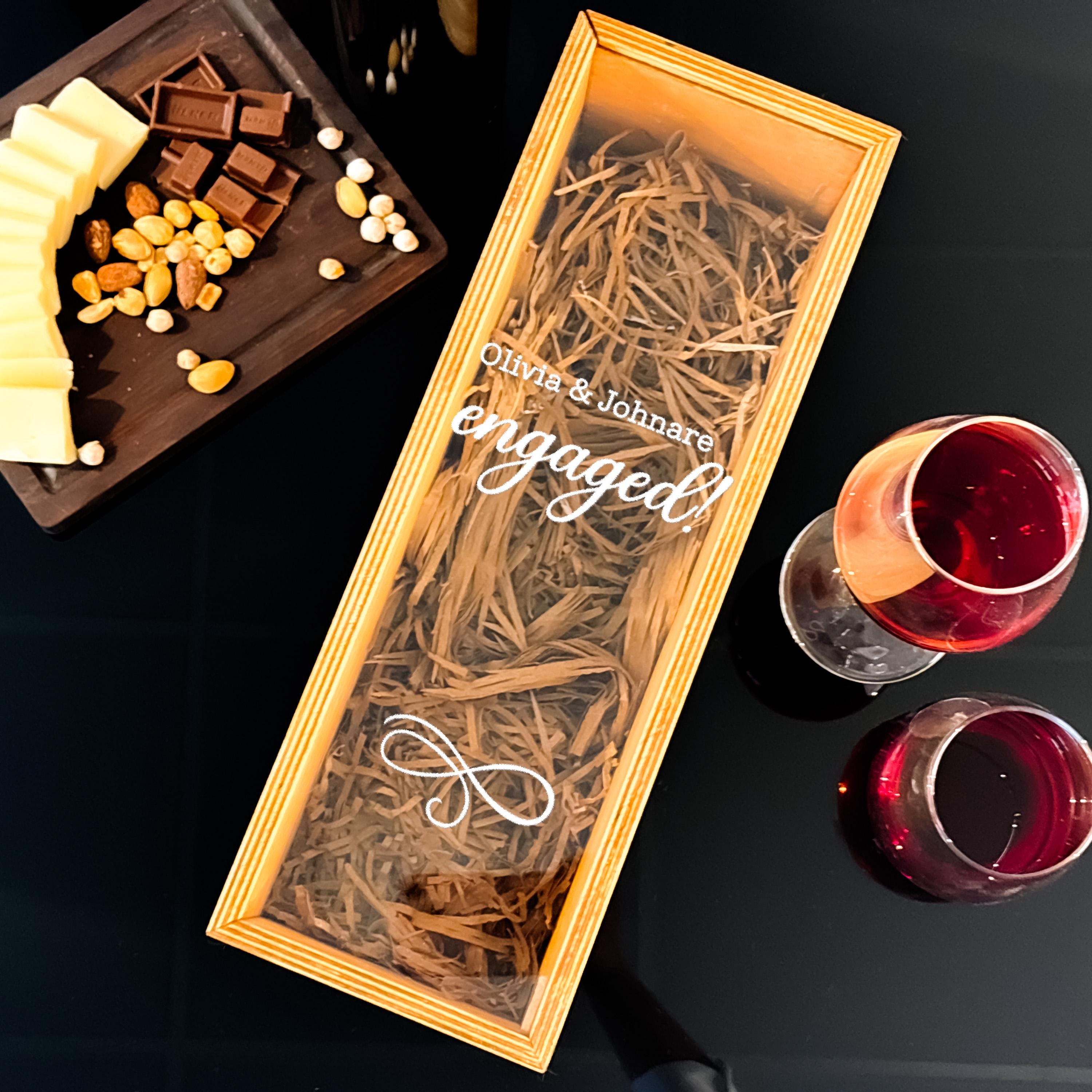 A wooden box with a clear lid, containing straw and a message, sits on a table next to a plate of cheese and chocolate. There are also two glasses of red wine and a small plate of nuts.