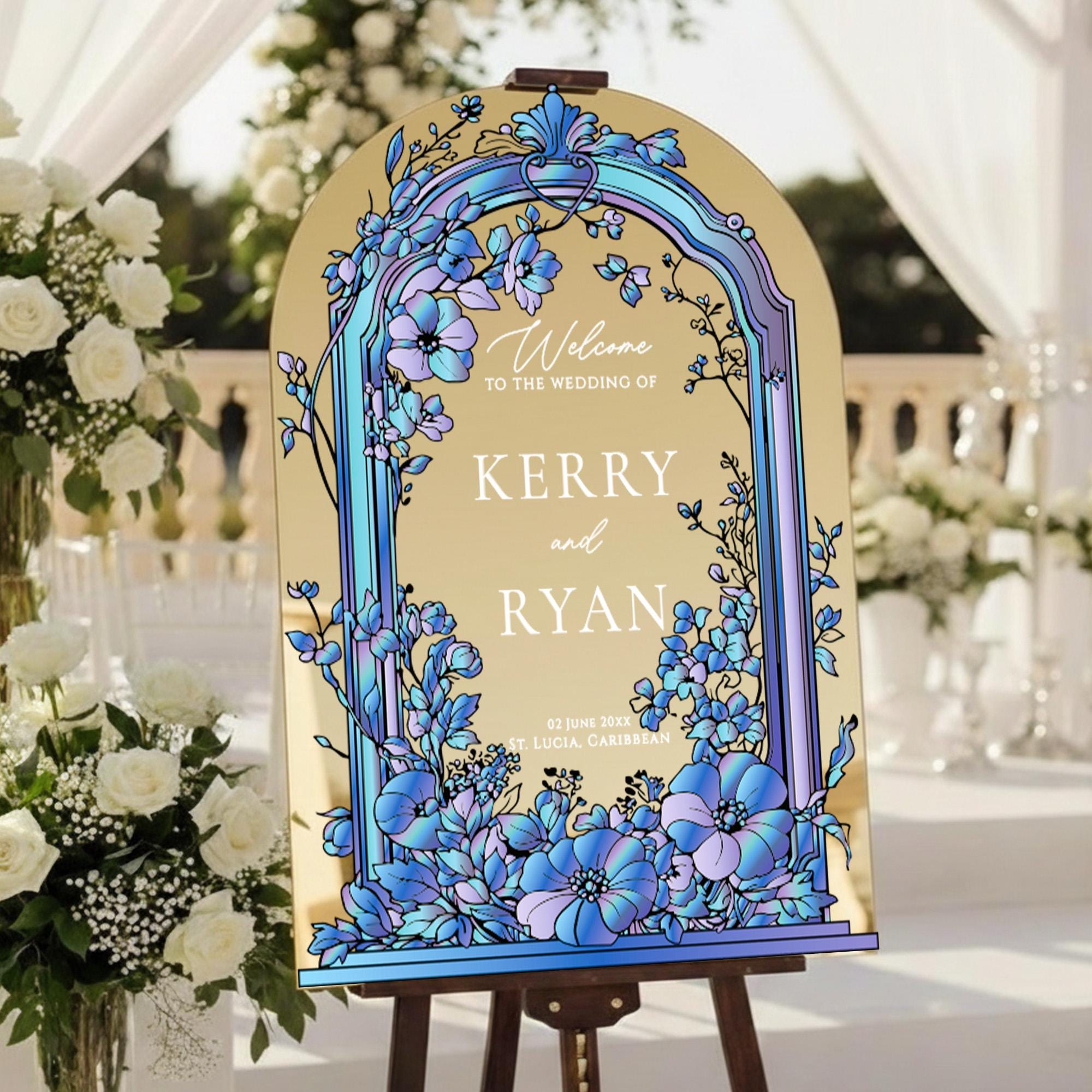 A wedding welcome sign with a floral design and the names 'Kerry' and 'Ryan' on it, placed on a wooden stand in a garden setting with white flowers and a white canopy in the background.