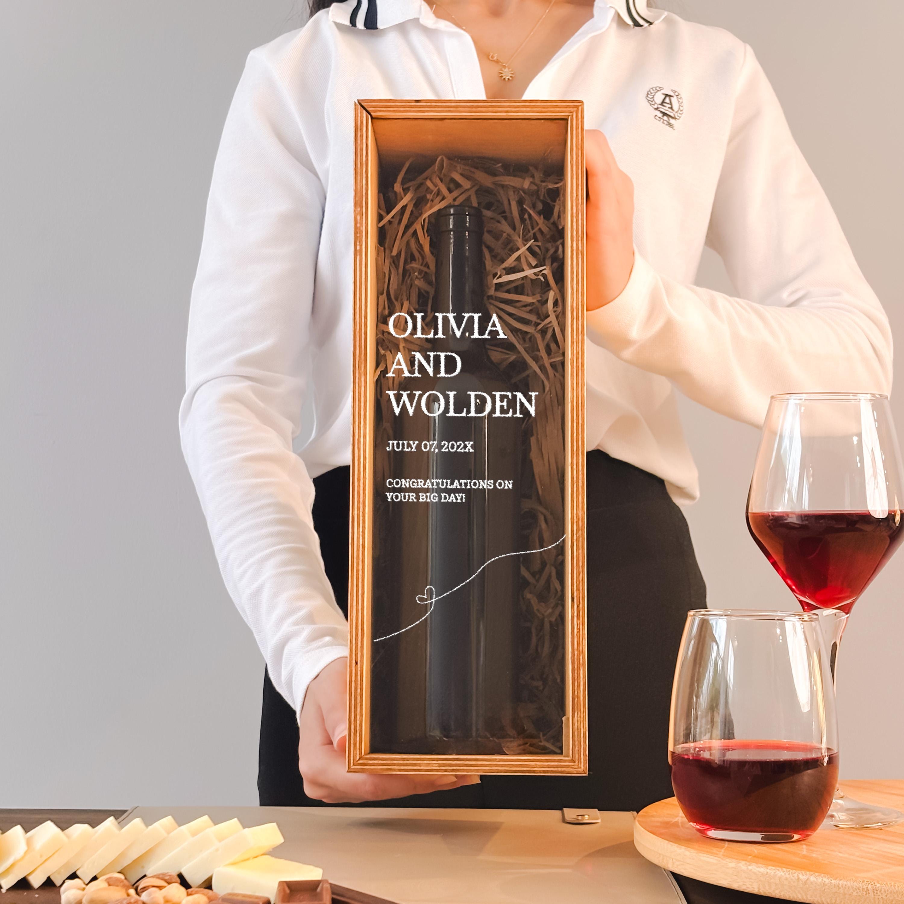 A person is holding a wooden box containing a bottle of wine, with a glass of red wine next to it.