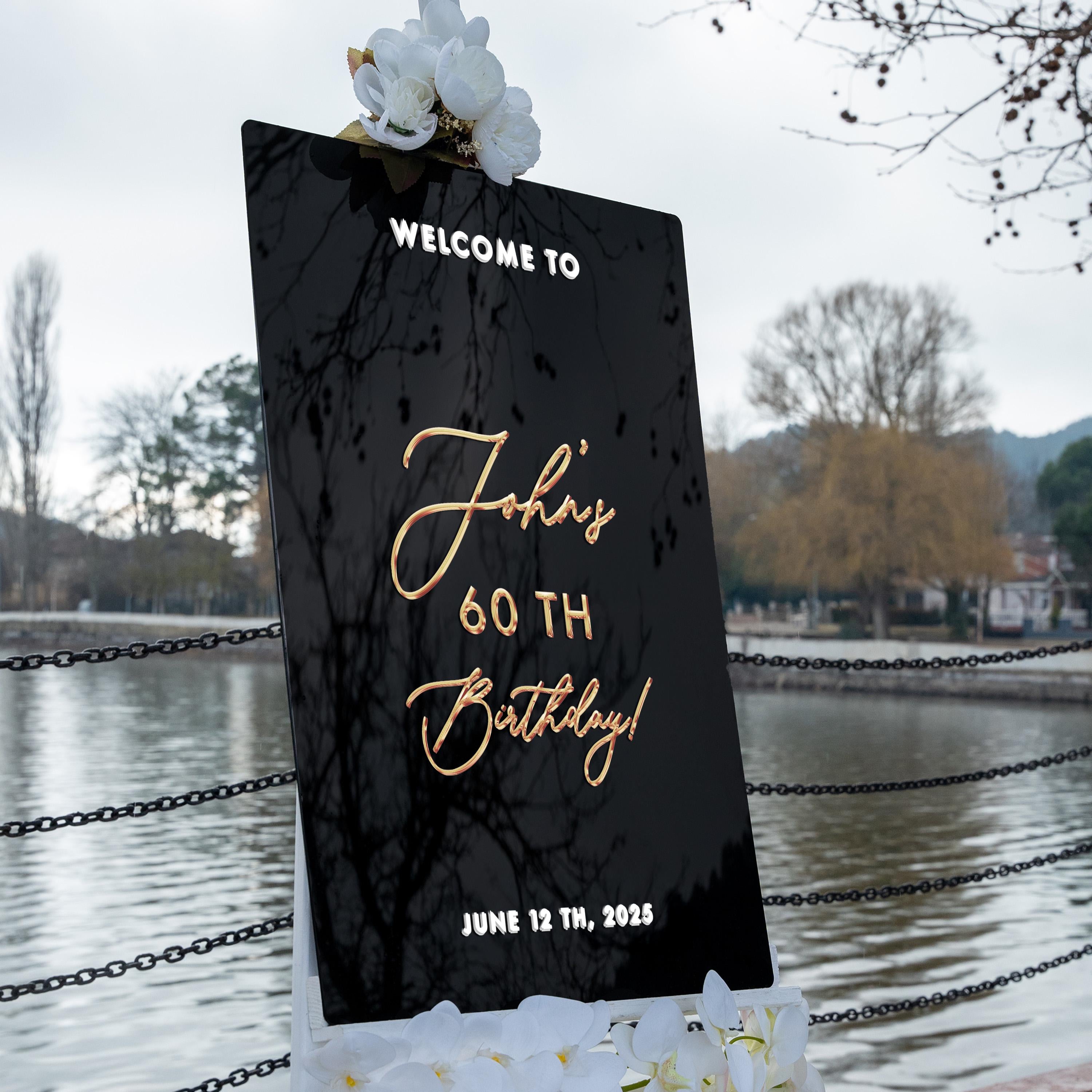 a black sign with gold text that reads "Welcome to John's 60th Birthday" and "June 12th, 2025". The sign is placed near a body of water, with trees and a fence in the background.