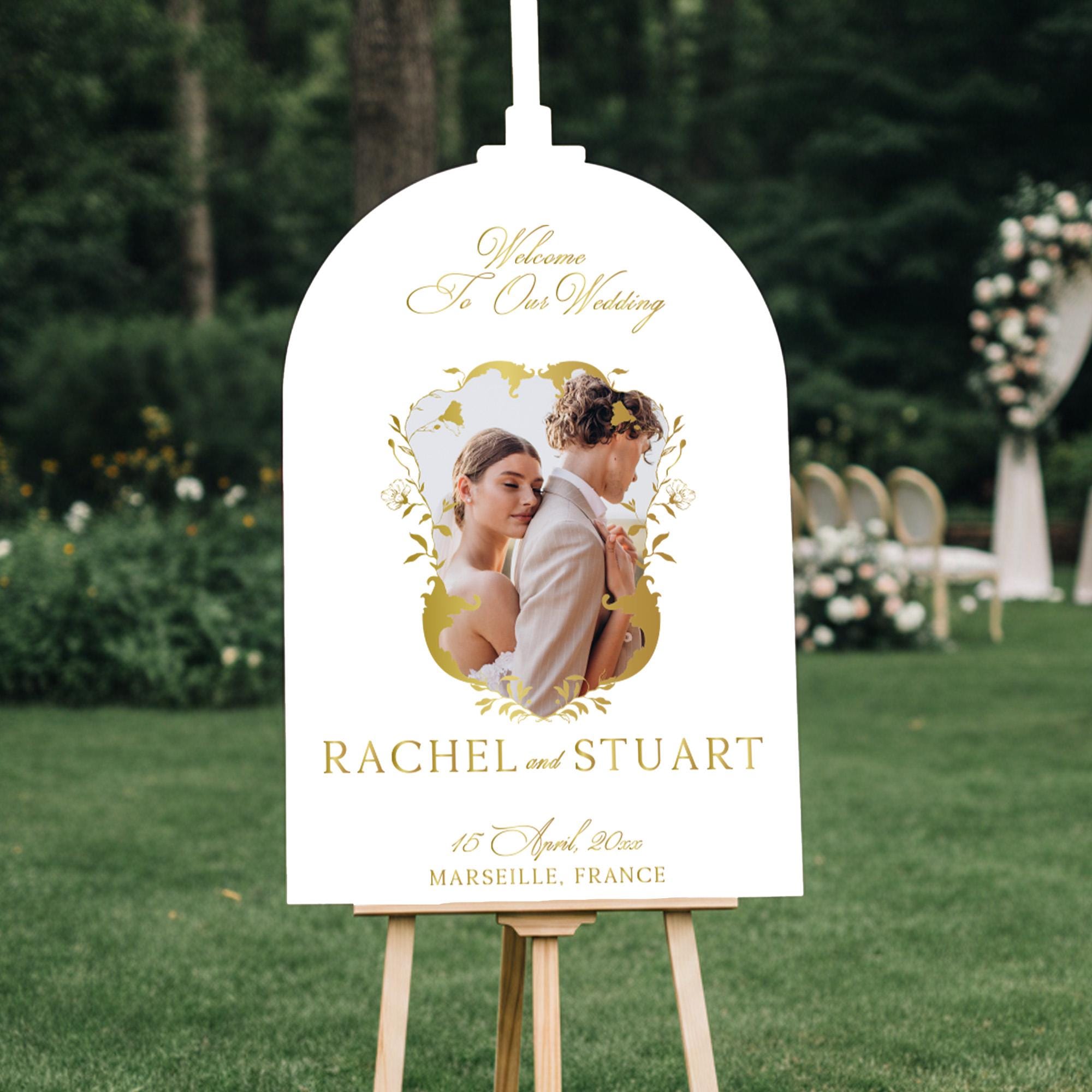 a white sign with a gold border, displaying a photo of a couple embracing. The sign reads "Welcome To Our Wedding" and "RACHEL and STUART" along with the date "15 April, 2020 MARSEILLE, FRANCE".