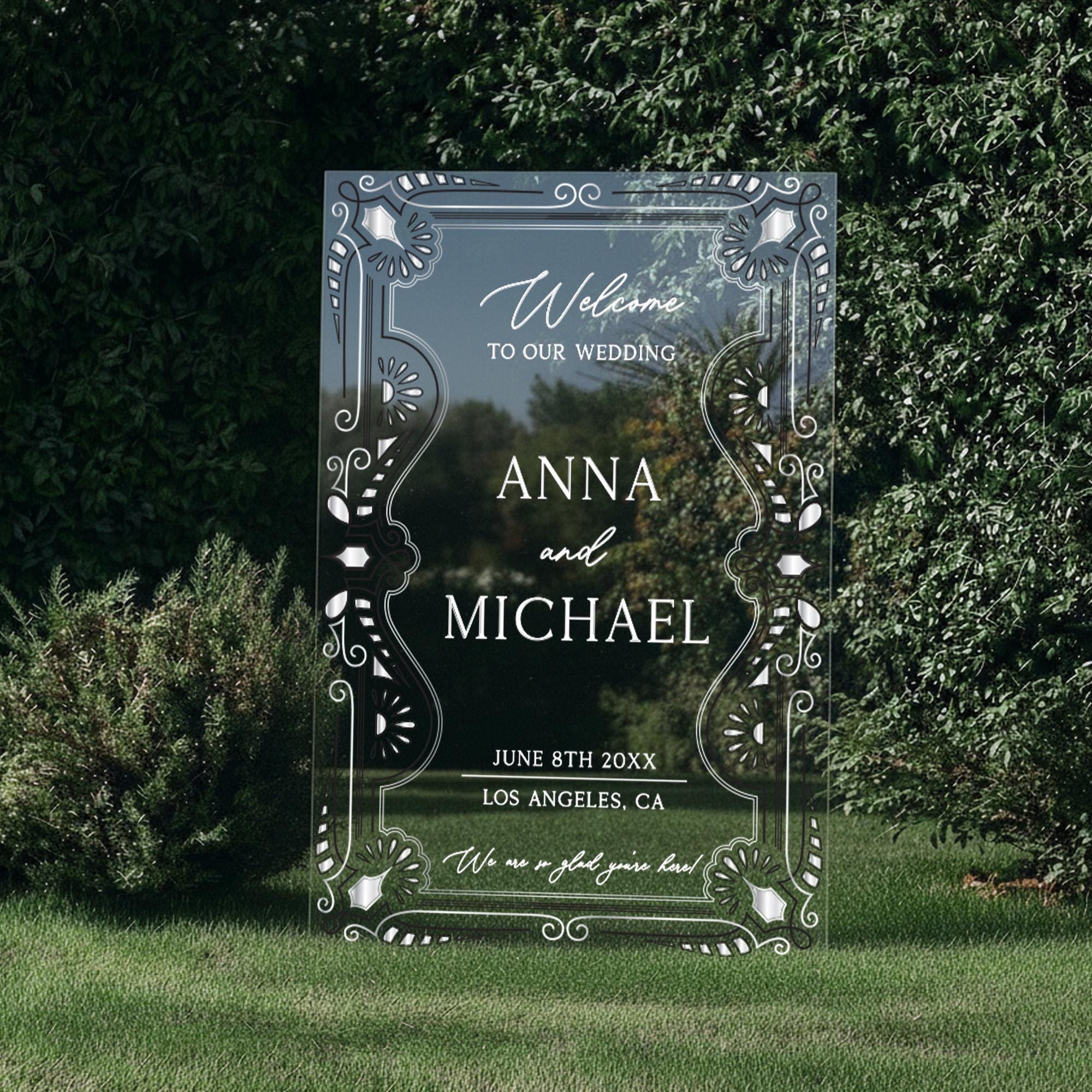 a large, ornate, and decorative wedding invitation sign with the names 'Anna and Michael' on it, placed in a grassy area surrounded by greenery.