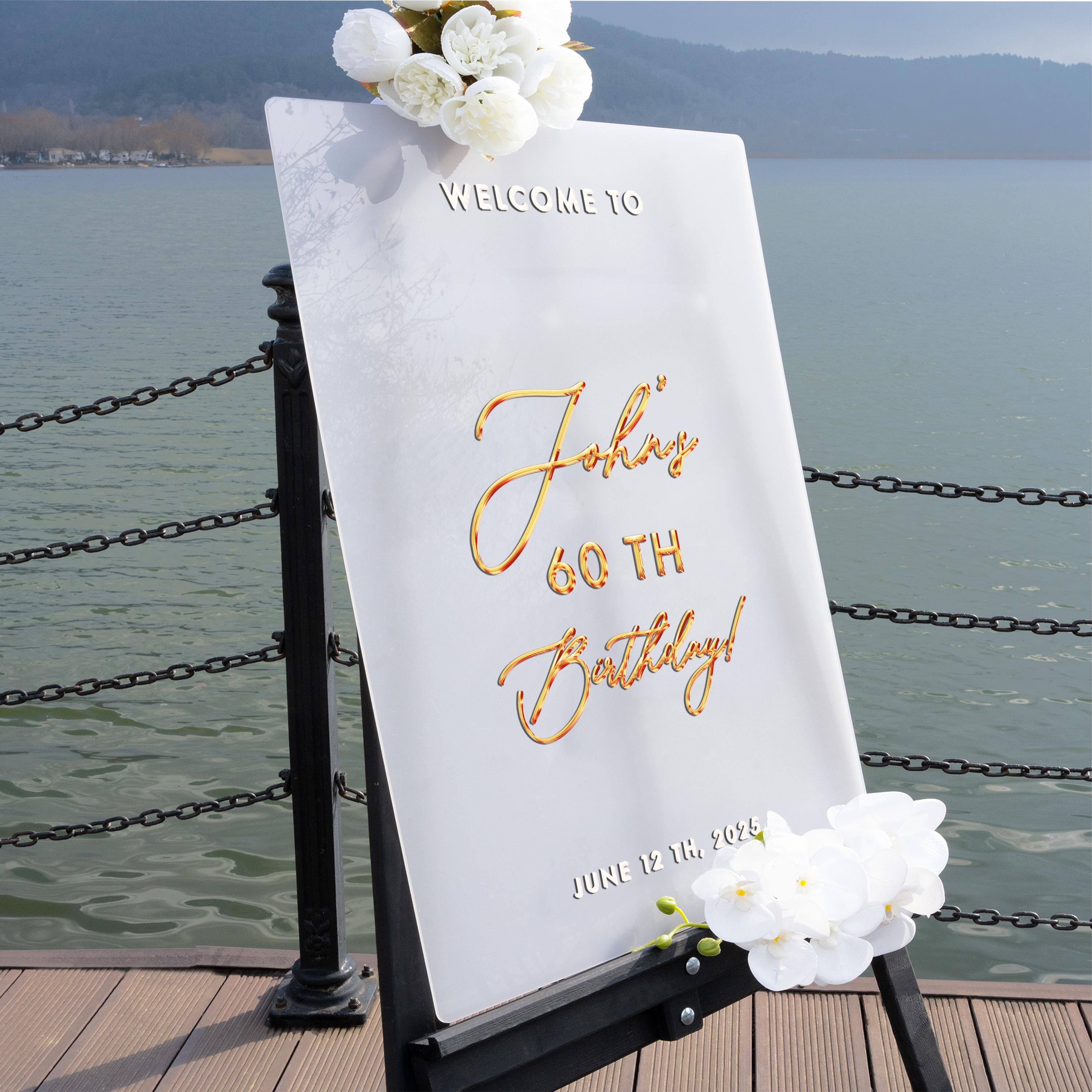 A white sign with gold lettering welcoming guests to a 60th birthday celebration, placed on a wooden dock overlooking a body of water.