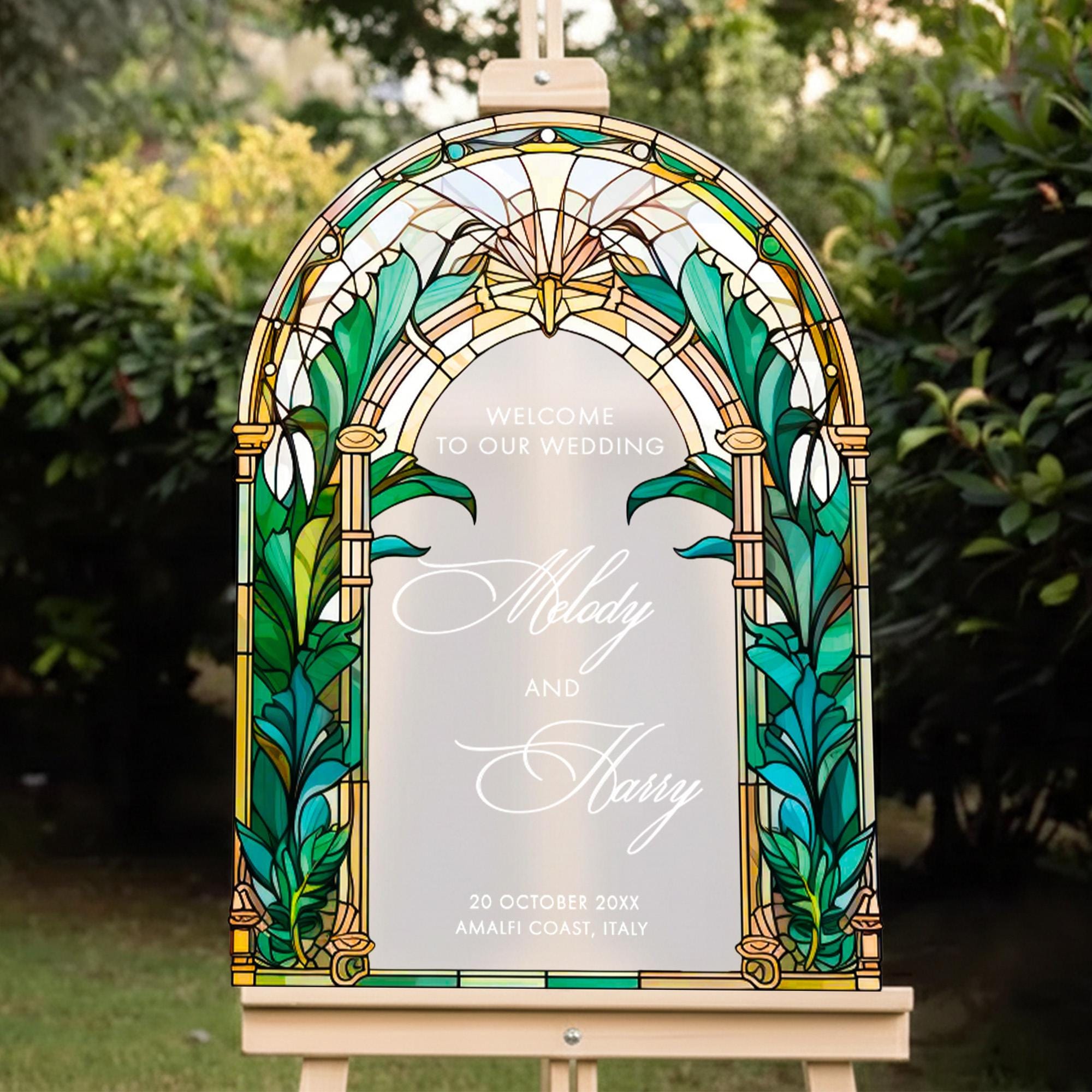 A stained glass-style wedding sign with the names 'Melody and Harry' and the date '20 October 20xx' displayed on it, set against a backdrop of greenery.