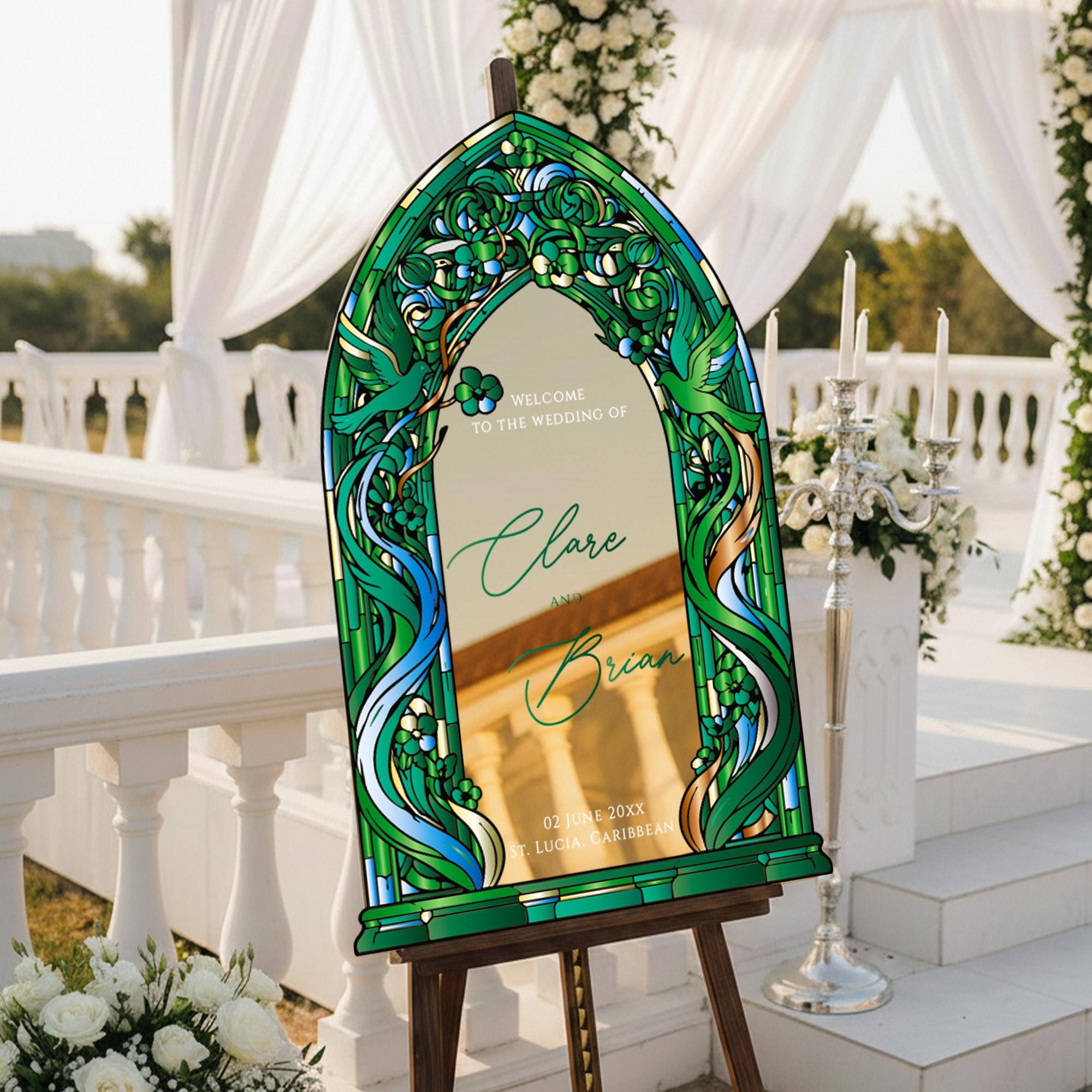 A wedding invitation sign with a green and gold design, featuring the names 'Clare' and 'Brian', stands on a wooden stand in front of a white archway decorated with flowers.