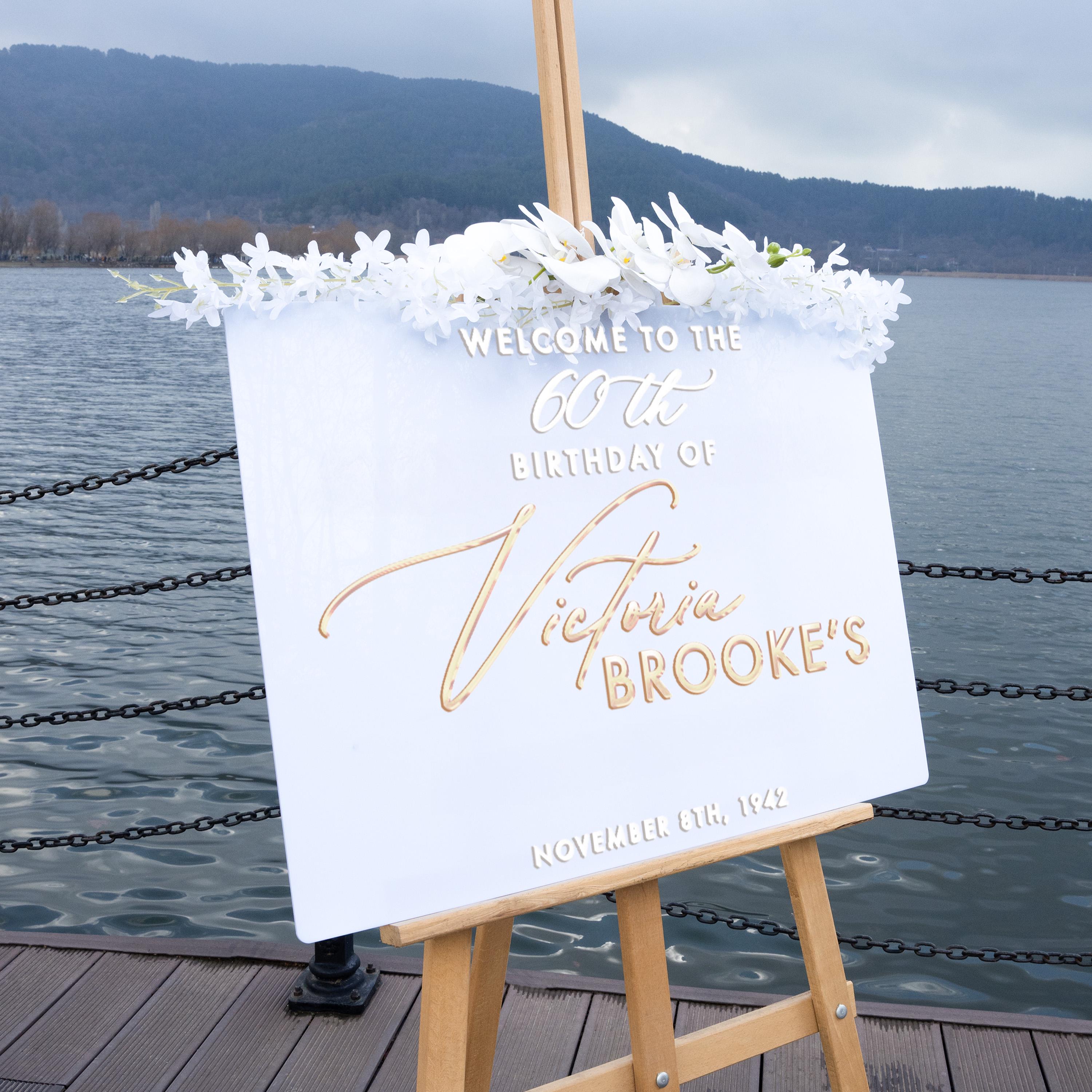 a white sign with gold lettering that reads "Welcome to the 60th Birthday of Victoria Brooke's" and "NOVEMBER 8TH, 1942" below it. The sign is placed on a wooden dock overlooking a body of water, with mountains in the background.
