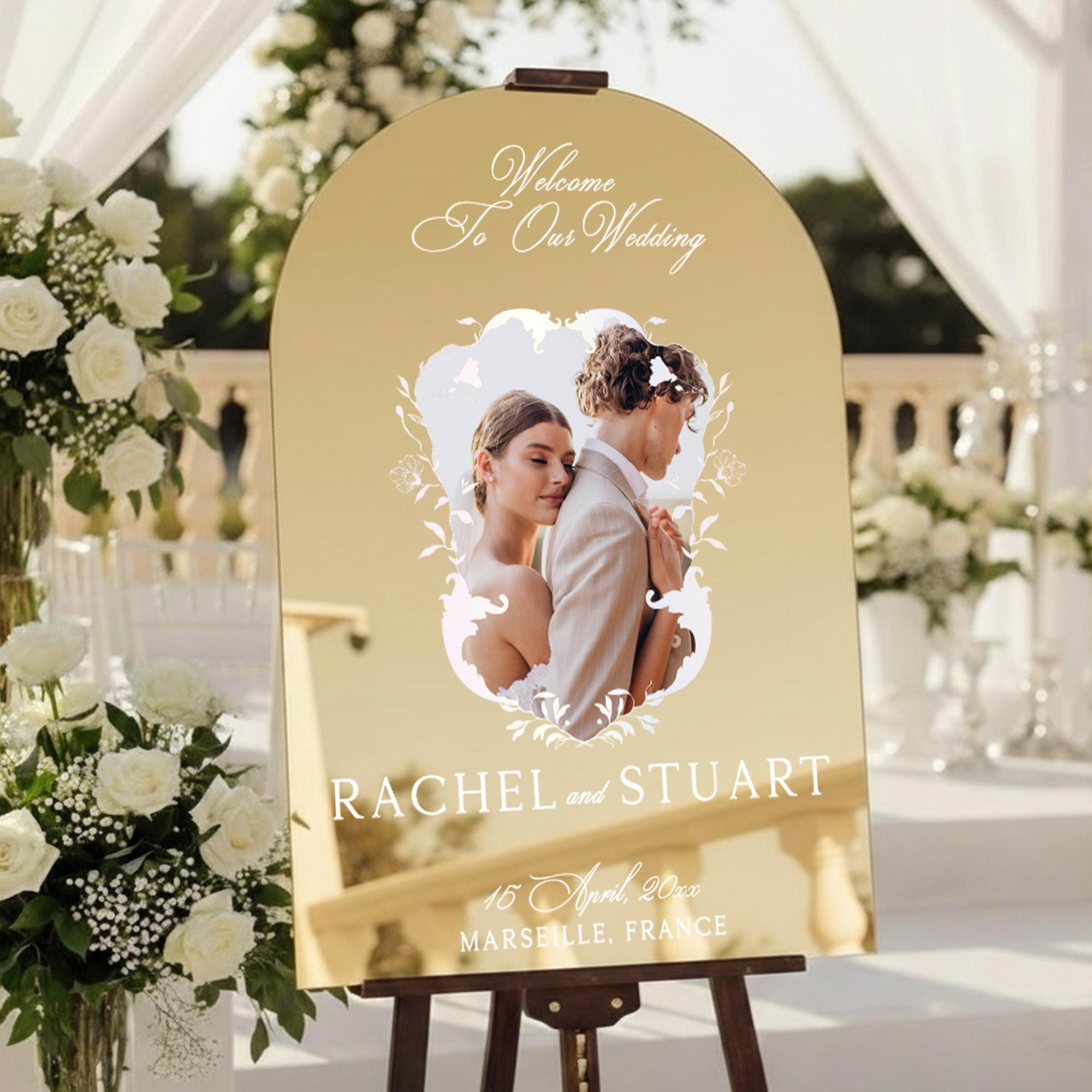 a wedding invitation sign with a couple in a romantic embrace, surrounded by white flowers and greenery. The sign reads "Welcome To Our Wedding" and "RACHEL and STUART" along with the date "16 April, 2020 MARSELLE, FRANCE".