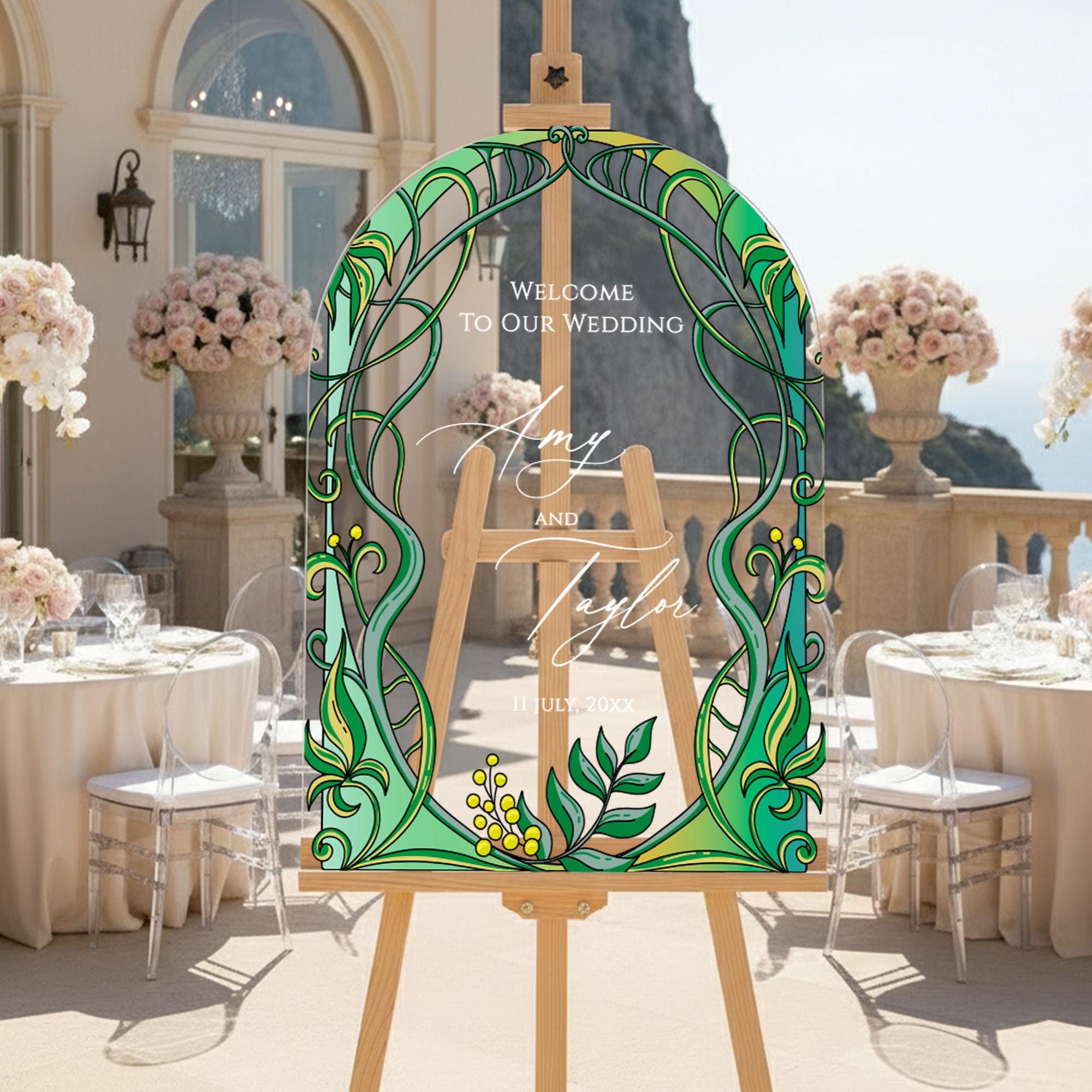 a wedding reception setup with a large, colorful, and ornate sign that reads "Welcome to our wedding Amy and Taylor July 20xx" in the foreground. The sign is surrounded by a table with white tablecloths, floral centerpieces, and clear plastic chairs. The background includes a balcony overlooking the ocean, adding to the scenic ambiance of the wedding celebration.