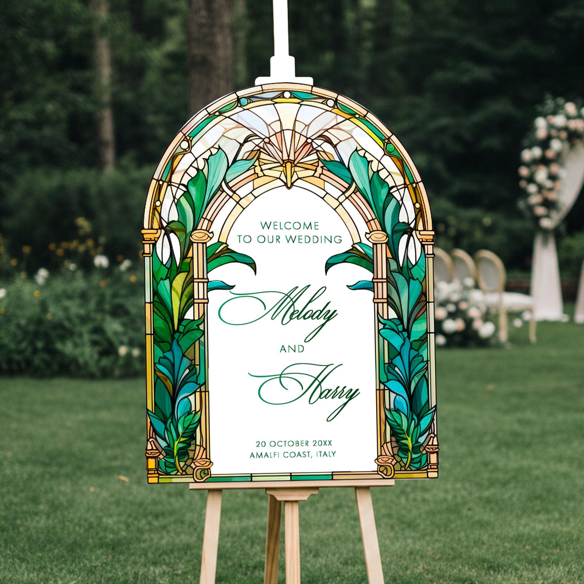 A stained glass-style wedding welcome sign with the names 'Melody' and 'Harry' on it, standing on a wooden stand in a grassy area with trees in the background.