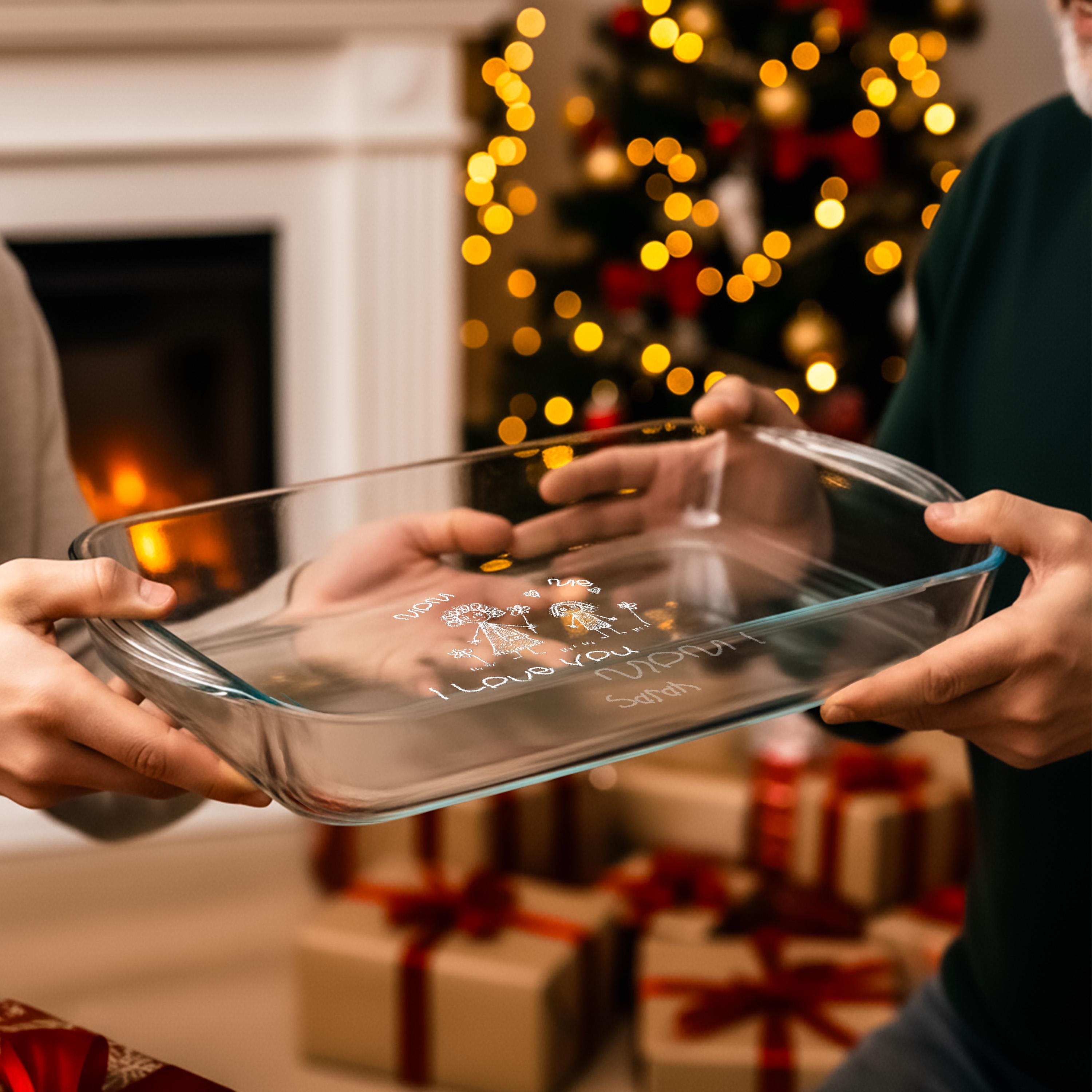 A person is holding a clear glass dish with a message written on it, in front of a Christmas tree with twinkling lights.