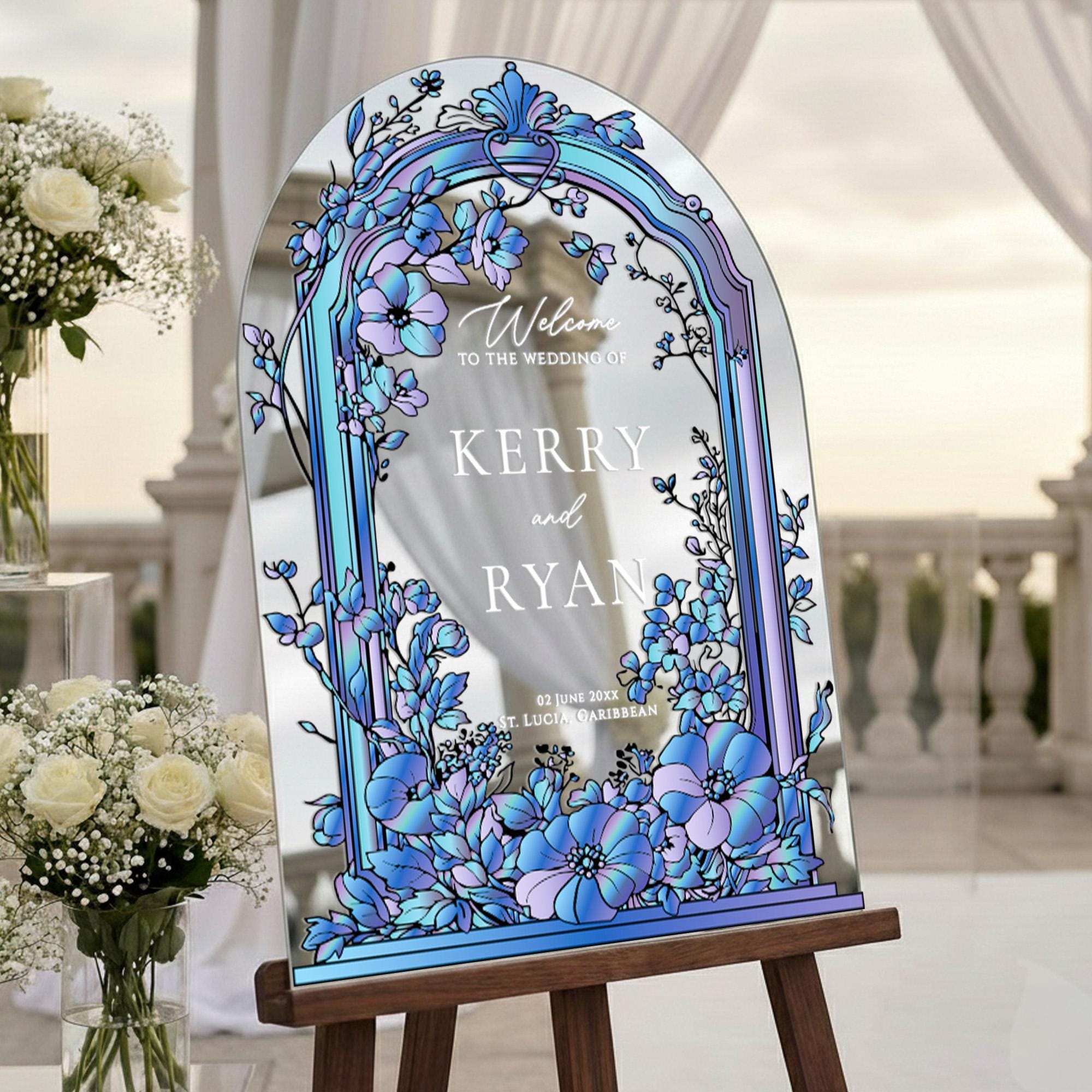 a large, ornate sign with a floral design, placed on a wooden stand. The sign reads "Welcome to the wedding of KERRY and RYAN" and is surrounded by a decorative arch. In the background, there are white flowers and a white curtain, creating a serene and elegant atmosphere.