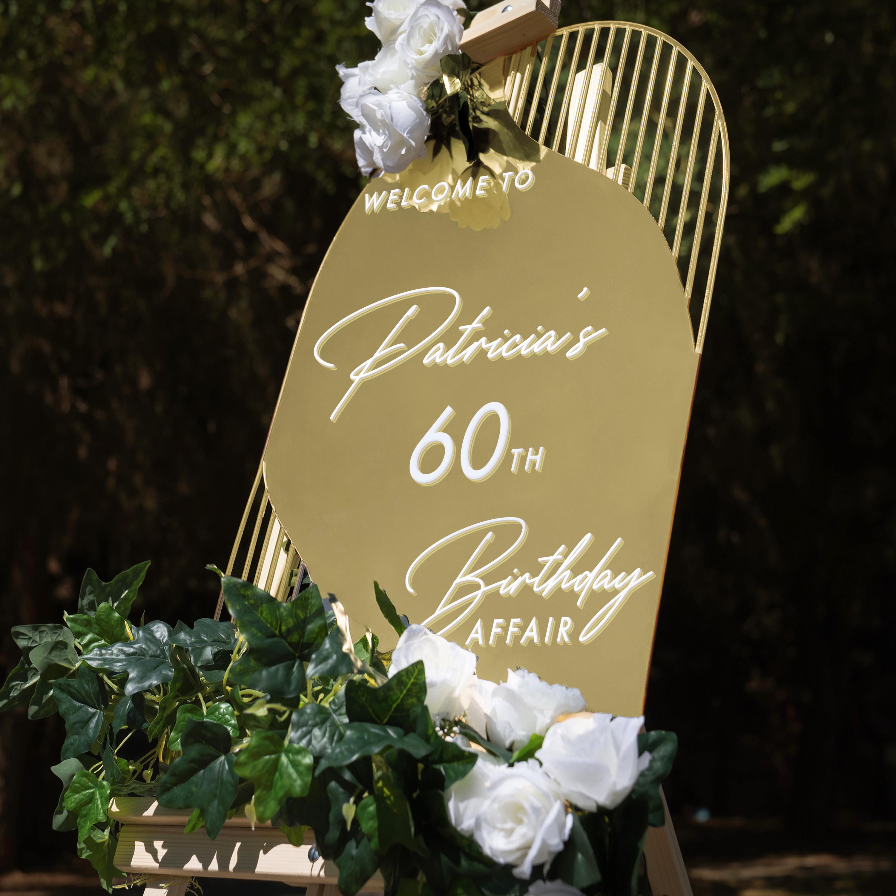 A golden sign welcoming guests to Patricia's 60th birthday party, with white flowers and greenery decorating the sign.