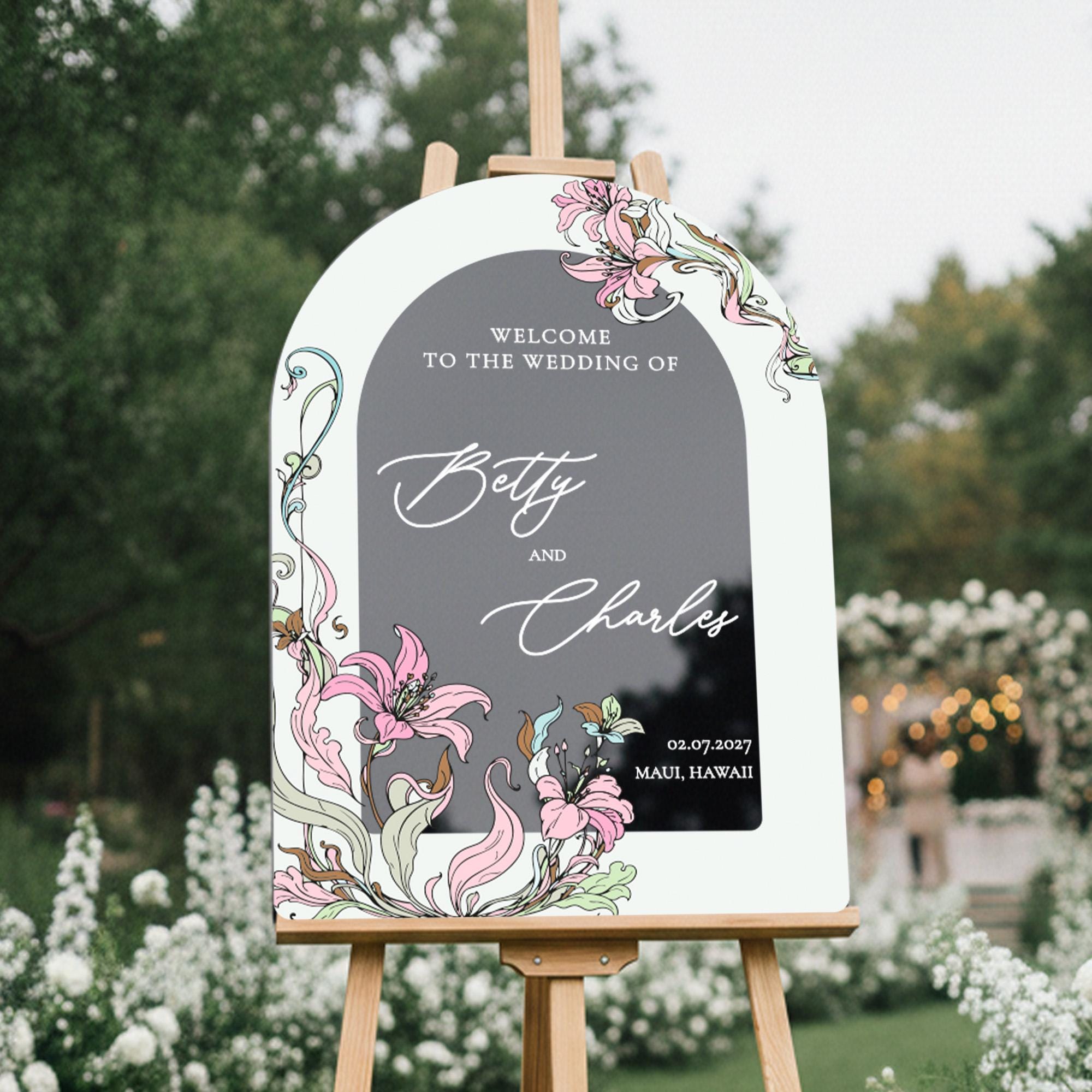 a white sign with floral designs and text, standing in a garden setting with trees and flowers in the background.