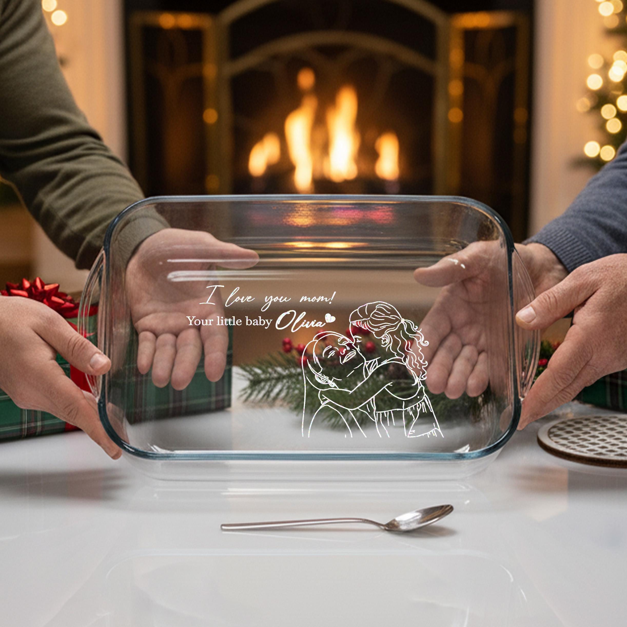 Two people holding a clear glass casserole dish with a personalized message on it, in front of a fireplace.