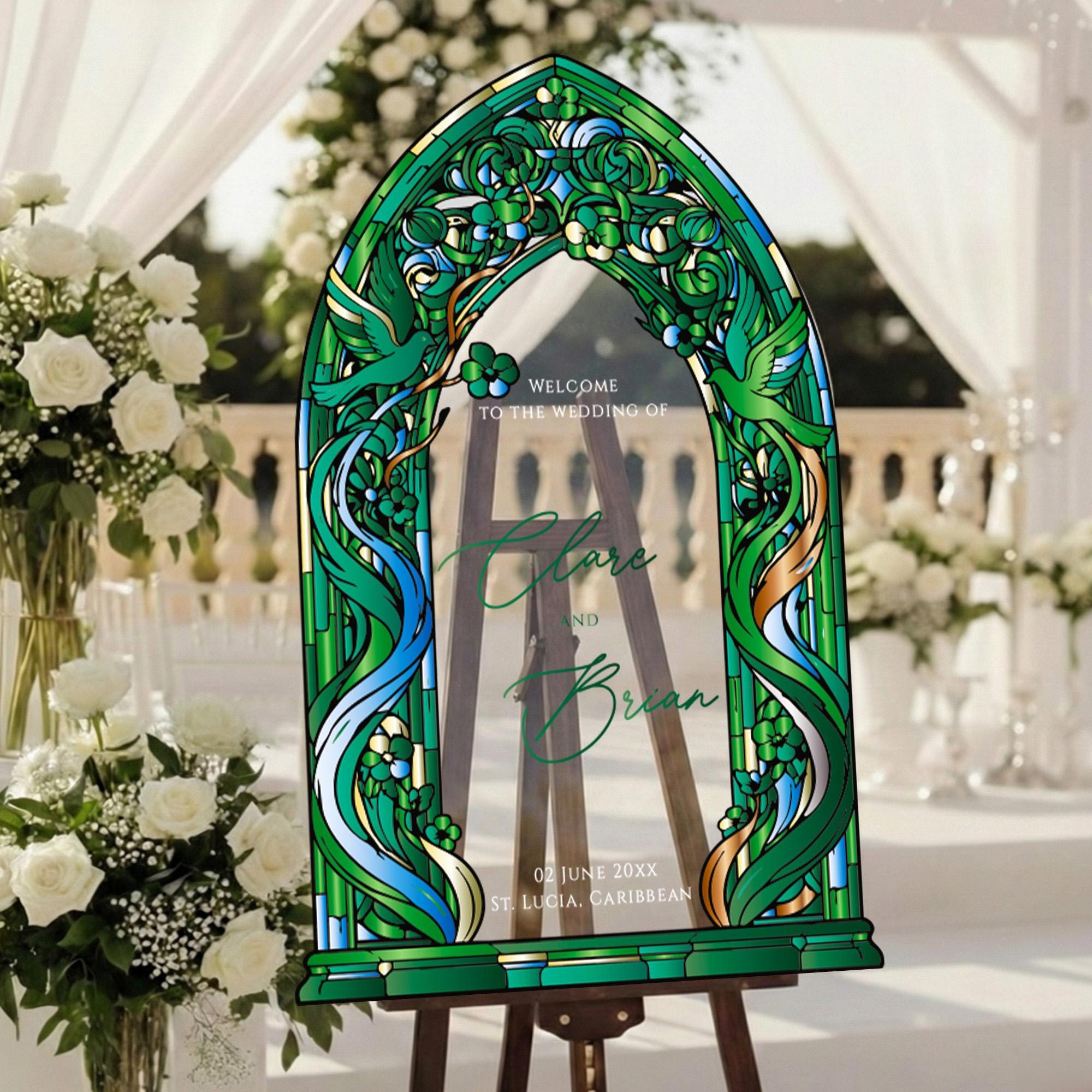 a large, ornate stained glass window with a green and blue design, placed in a garden setting with white flowers and greenery. The window has a welcoming message that reads "Welcome to the Wedding of Clare and Brian" and includes the date "02 JUNE 20XX ST. LUCIA CARIBBEAN".