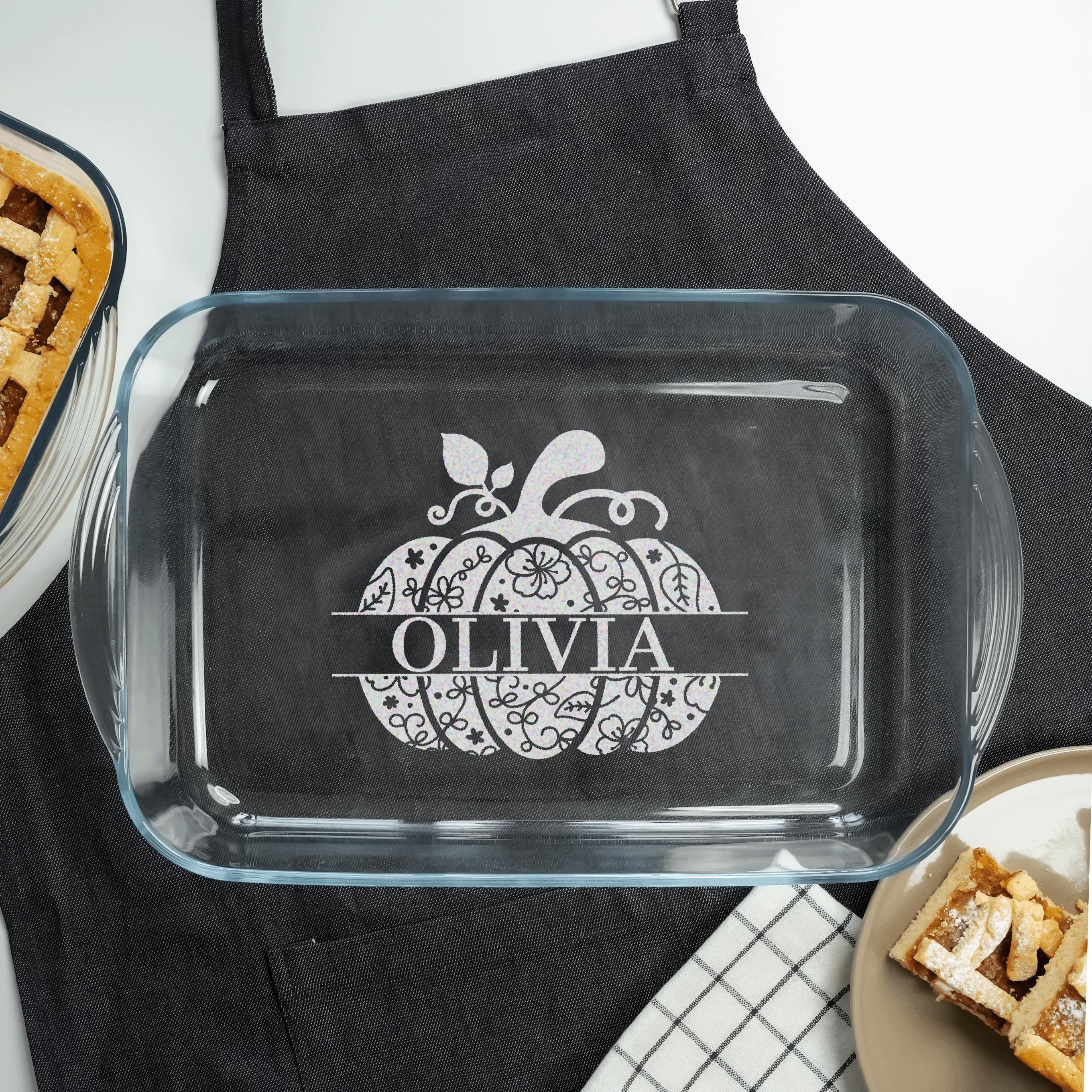 a clear glass casserole dish with a personalized pumpkin design and the name "OLIVIA" printed on it. The dish is placed on a table, and there is a black apron visible in the background.
