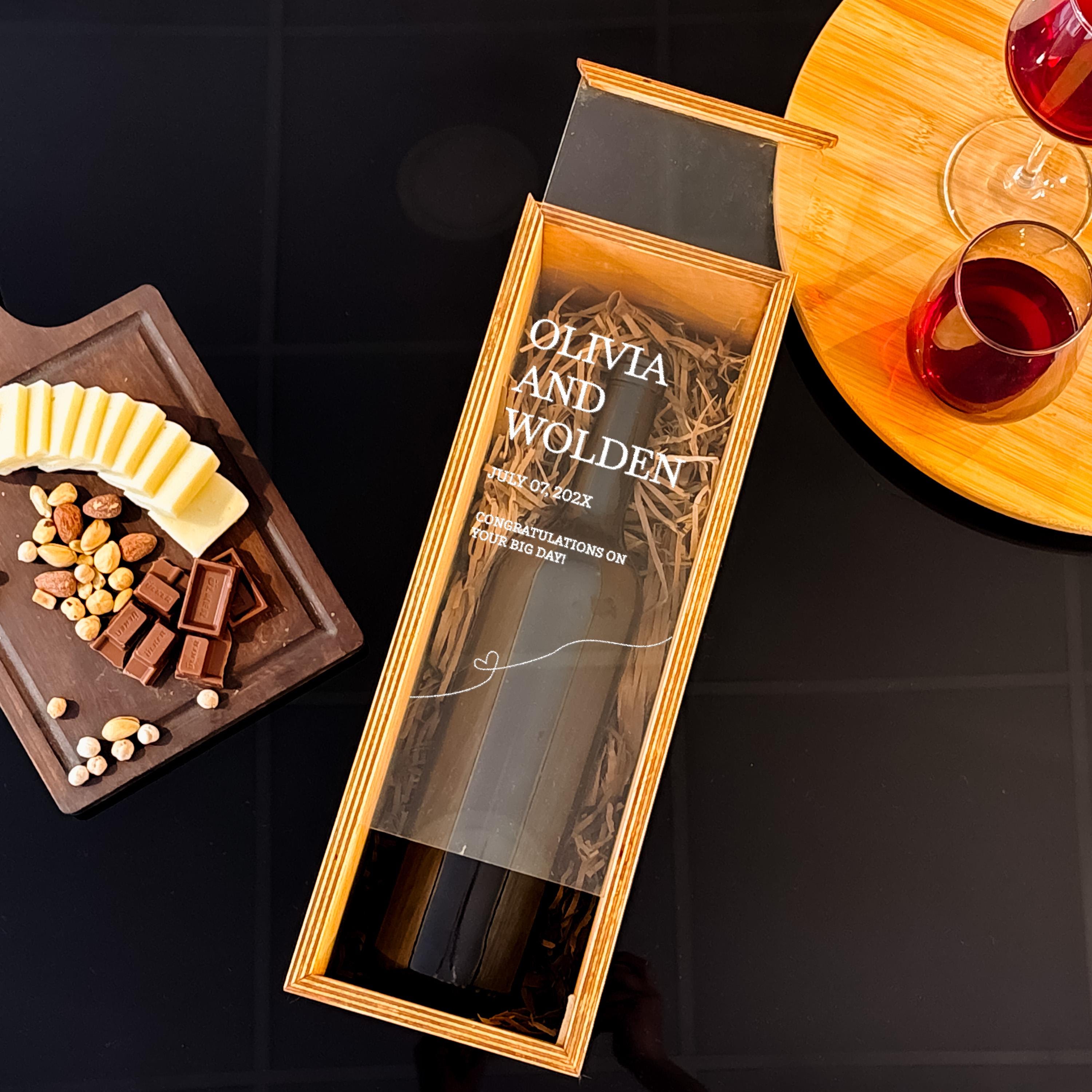 a wooden box with a clear plastic window, containing a bottle of wine, and a wooden tray with cheese and chocolate bars. There are also glasses of red wine and some nuts on the table.