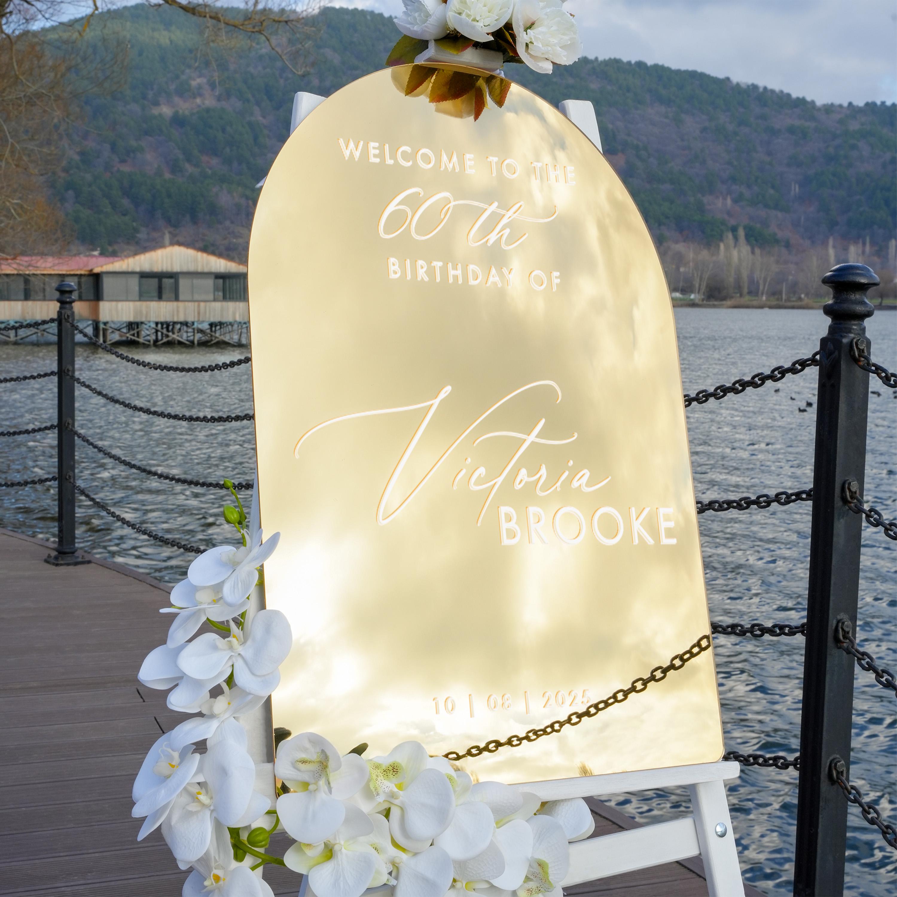 a golden sign that reads "Welcome to the 60th Birthday of Victoria Brooke" with a scenic background of a body of water and a bridge.