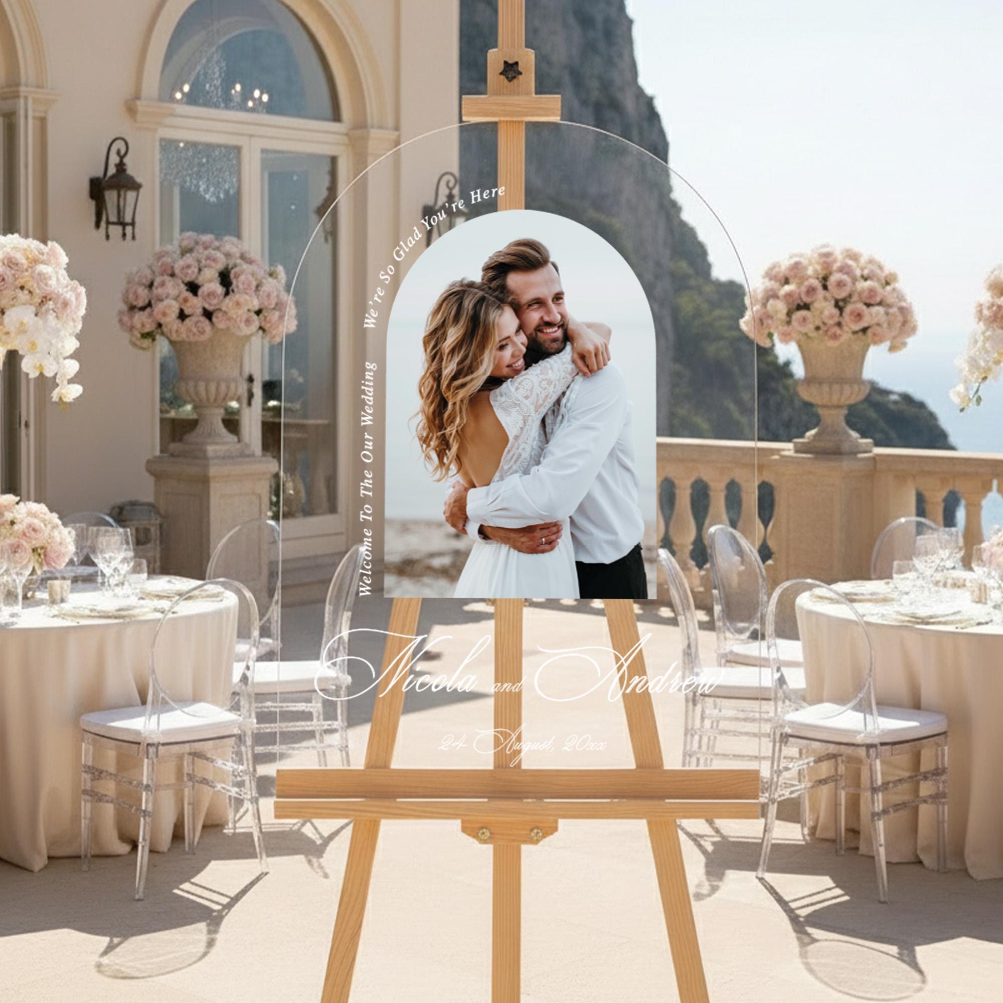 a wedding reception scene with a large wooden easel displaying a photo of a couple embracing. The couple is standing in front of a table set with white tablecloths and floral centerpieces. The background includes a scenic view of a cliff overlooking the ocean.