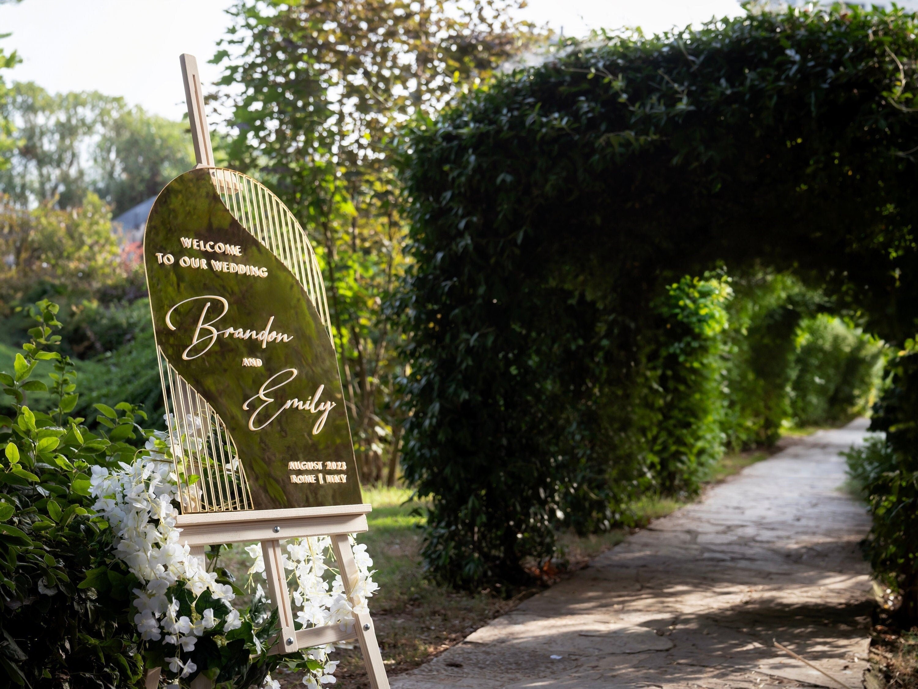 Gold mirror arch wedding welcome sign displayed on an easel at an outdoor wedding entrance, personalized with names, Rome Collection